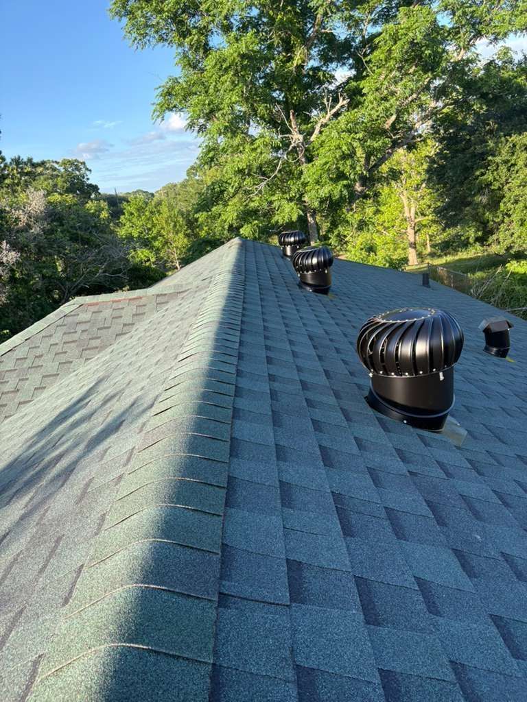 Roof with blue-gray shingles and black vents surrounded by green trees under a clear blue sky