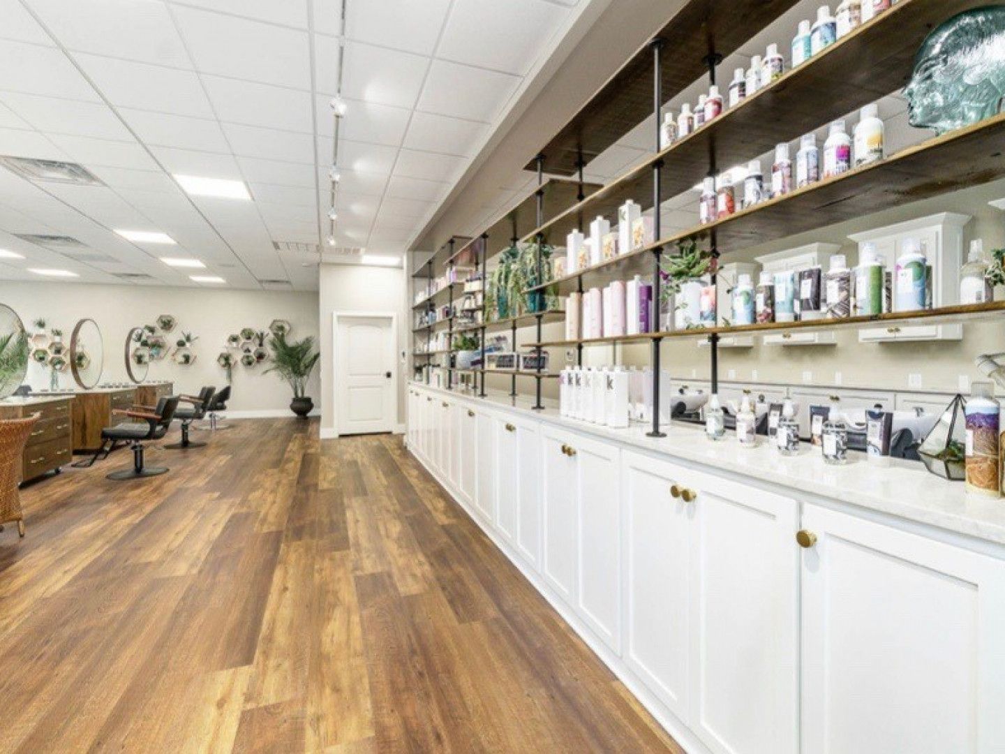 A hair salon with wooden floors , white cabinets and shelves filled with hair products.