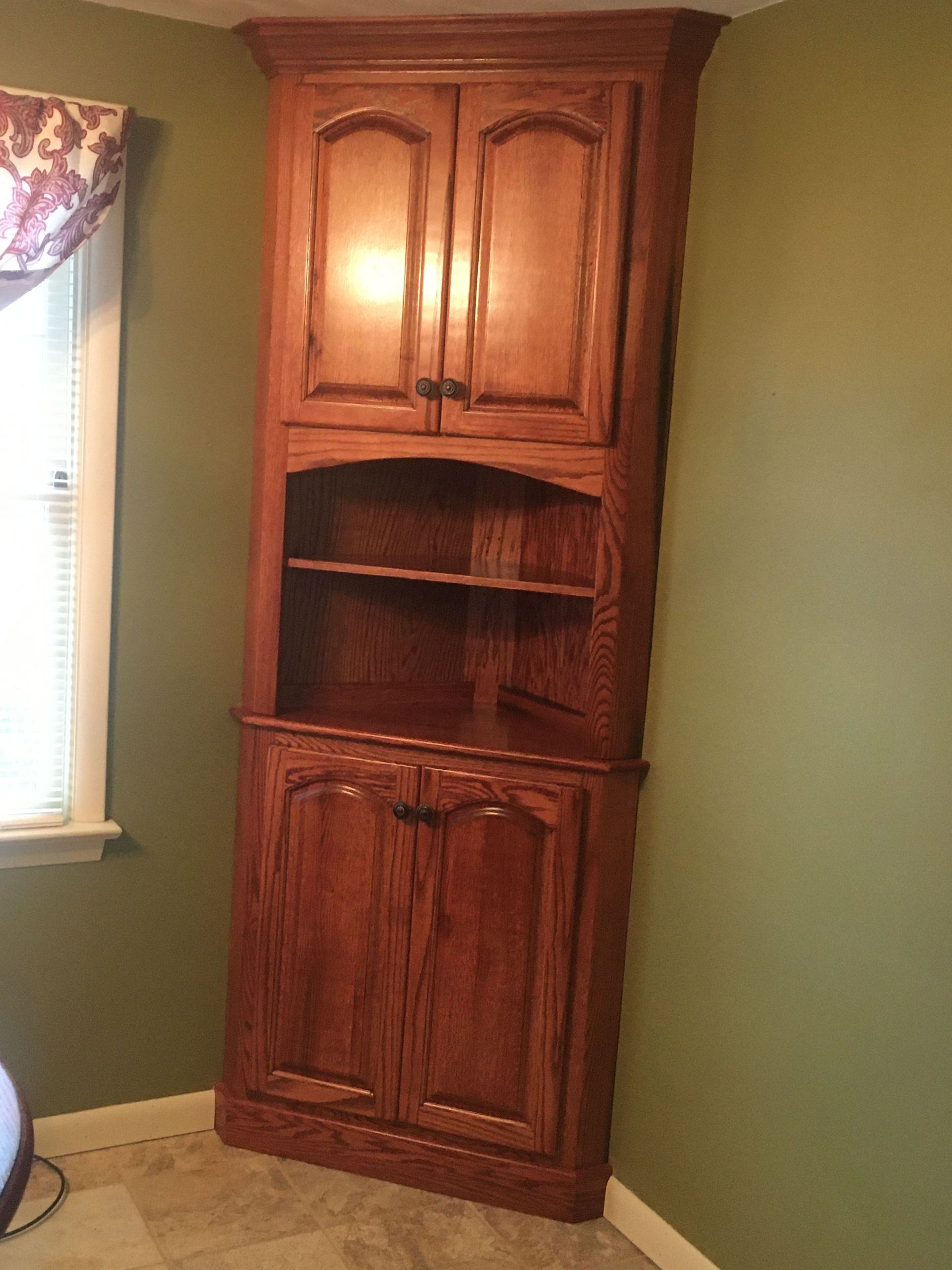 A wooden corner cabinet in a room next to a window.