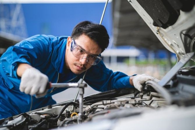 Mechanic in blue coveralls using a wrench under a car hood in a repair shop