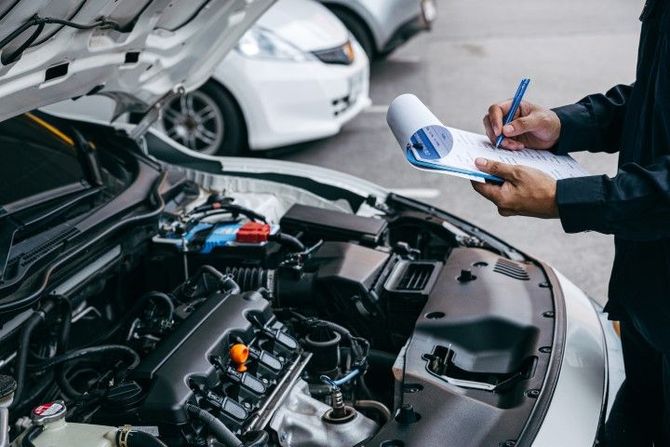 Mechanic inspecting a car engine and writing on a clipboard in an auto repair shop