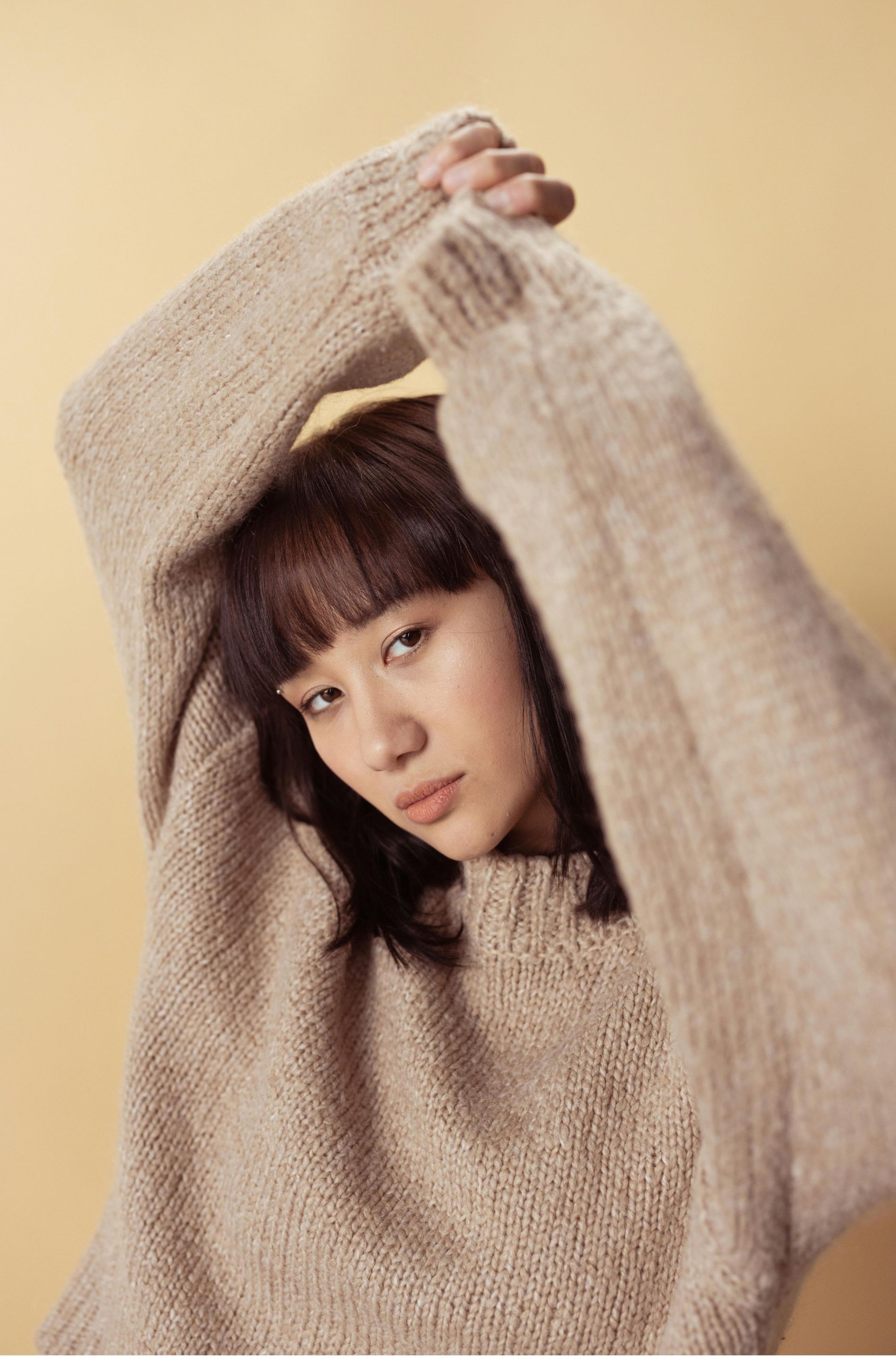 A woman is wearing a tan sweater with her arms over her head.