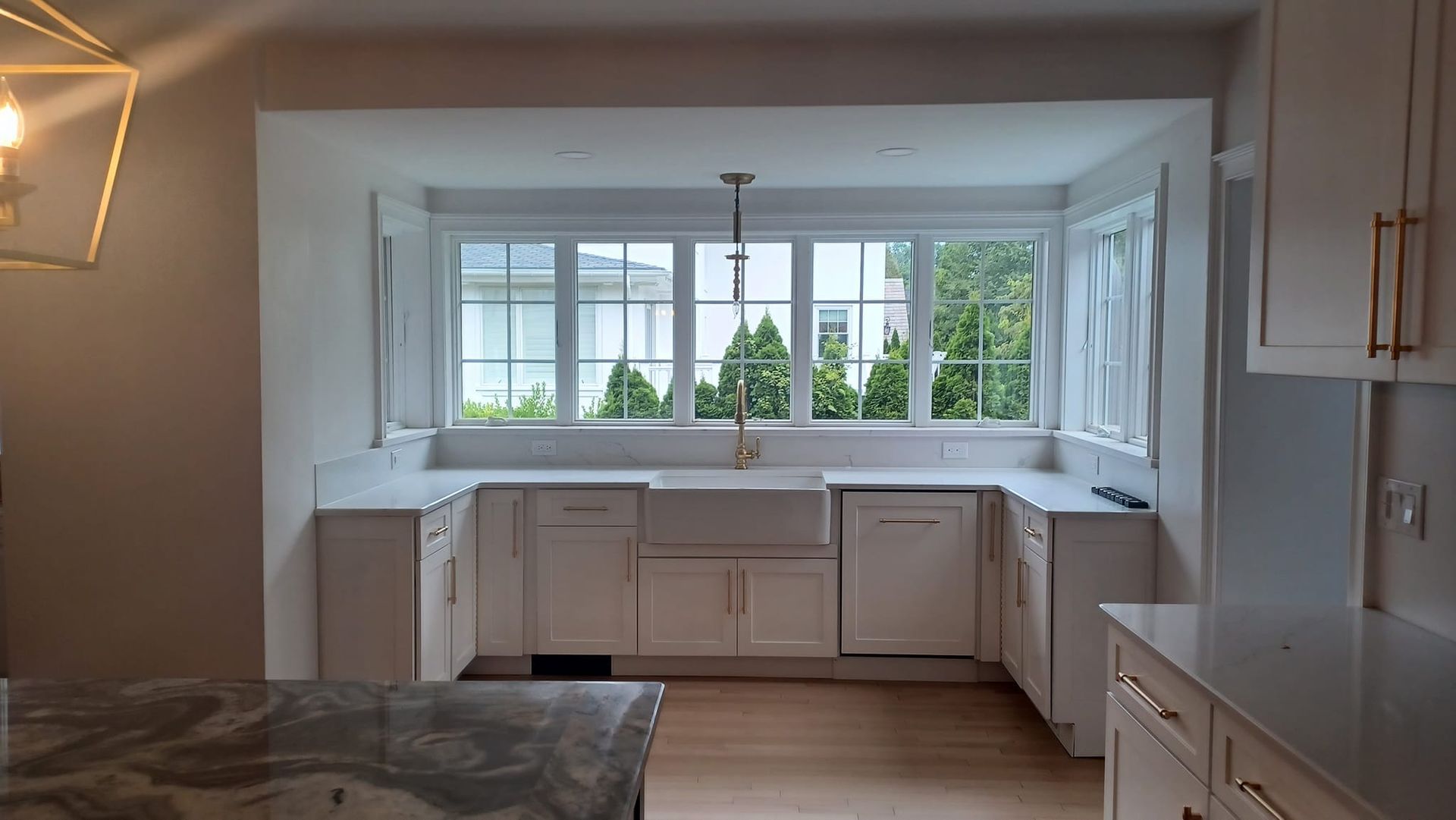 Bright kitchen with white cabinets, L-shaped counters, and large windows overlooking greenery.