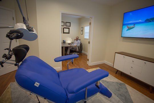 A blue massage chair in a light-colored room. A person works at a desk in the background, and a television displays a beach scene.