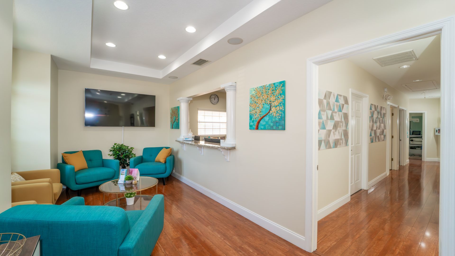 A waiting room with teal and yellow seating, a TV, and art on the walls. The hallway leads to treatment rooms.
