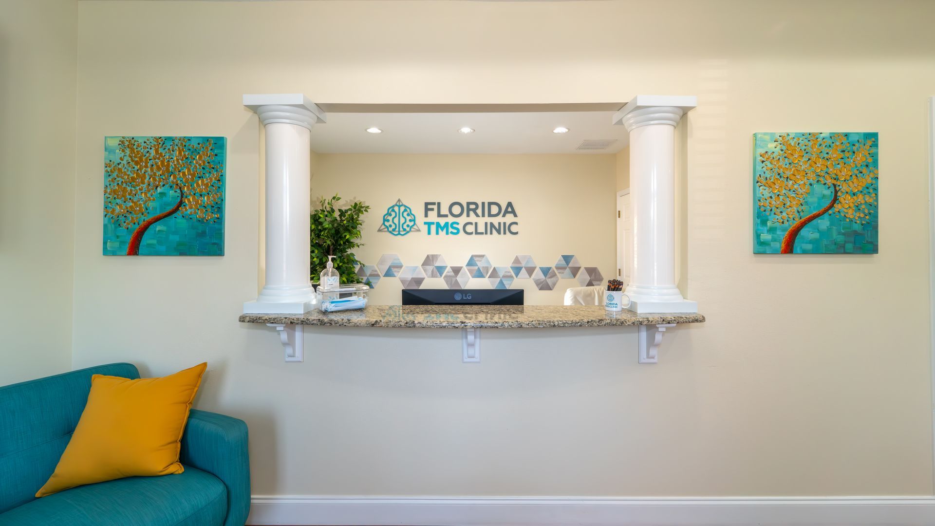 Reception area of Florida TMS Clinic. Turquoise sofa with yellow pillow. Desk area with logo, decorative accents, and artwork.
