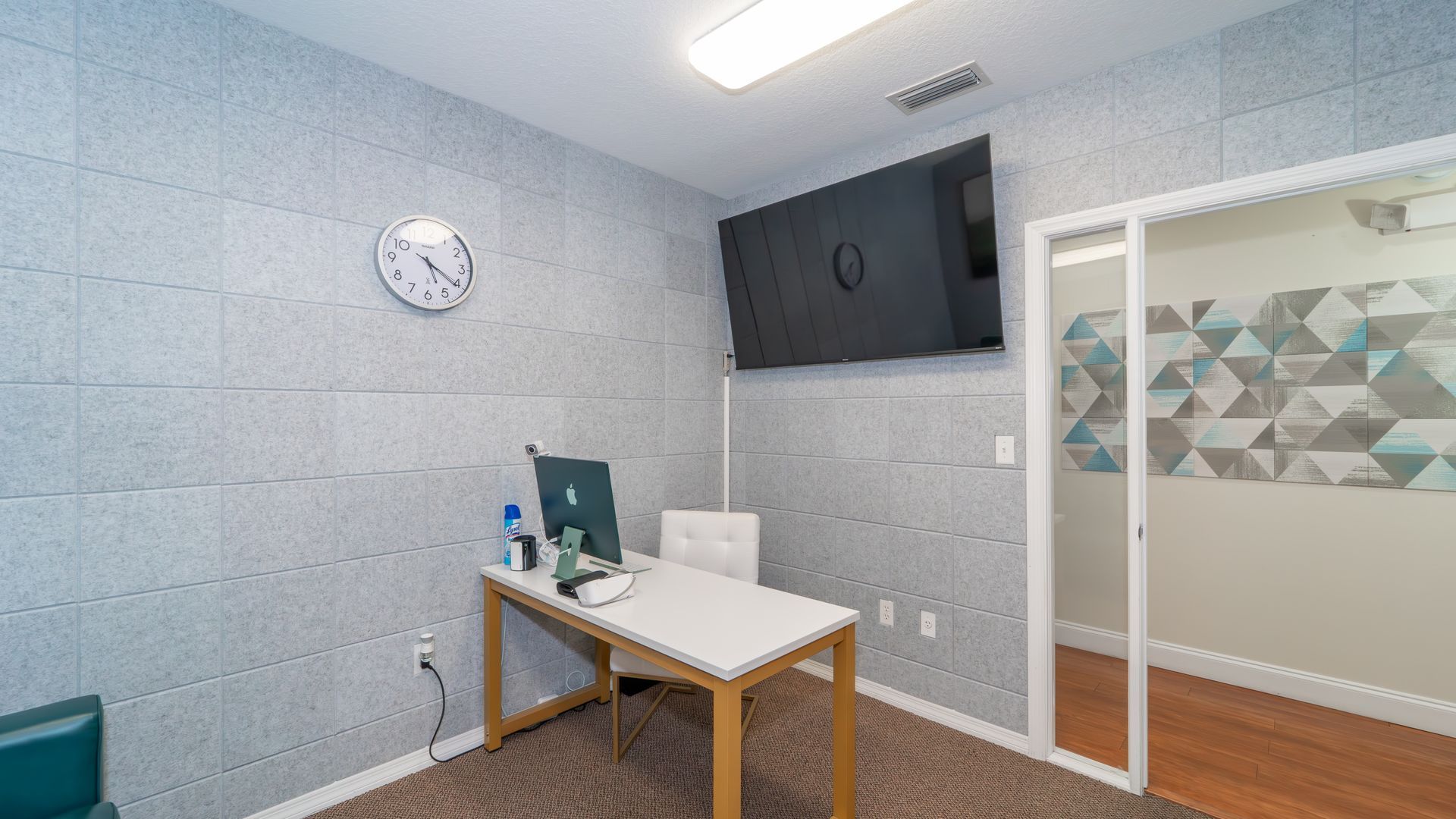 Office interior with a desk, computer, TV, and clock on the wall. A door leads to another room with geometric designs.
