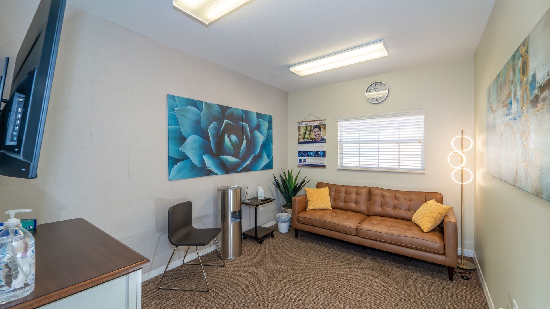 Cozy office waiting room. Brown leather sofa with yellow pillows, blue flower artwork, floor lamp, and desk with TV.