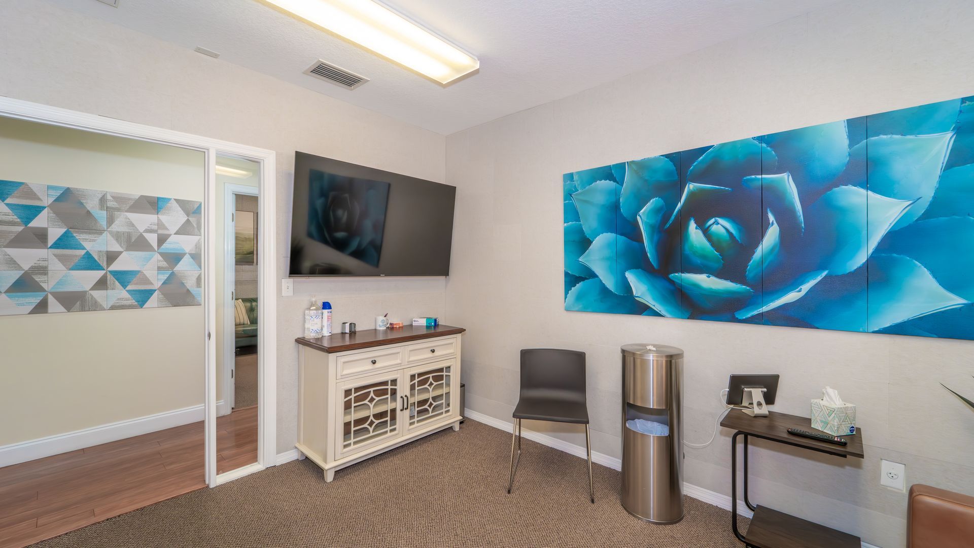 A waiting room with a large blue floral art piece. There's a TV above a cabinet, two chairs, and a water cooler.