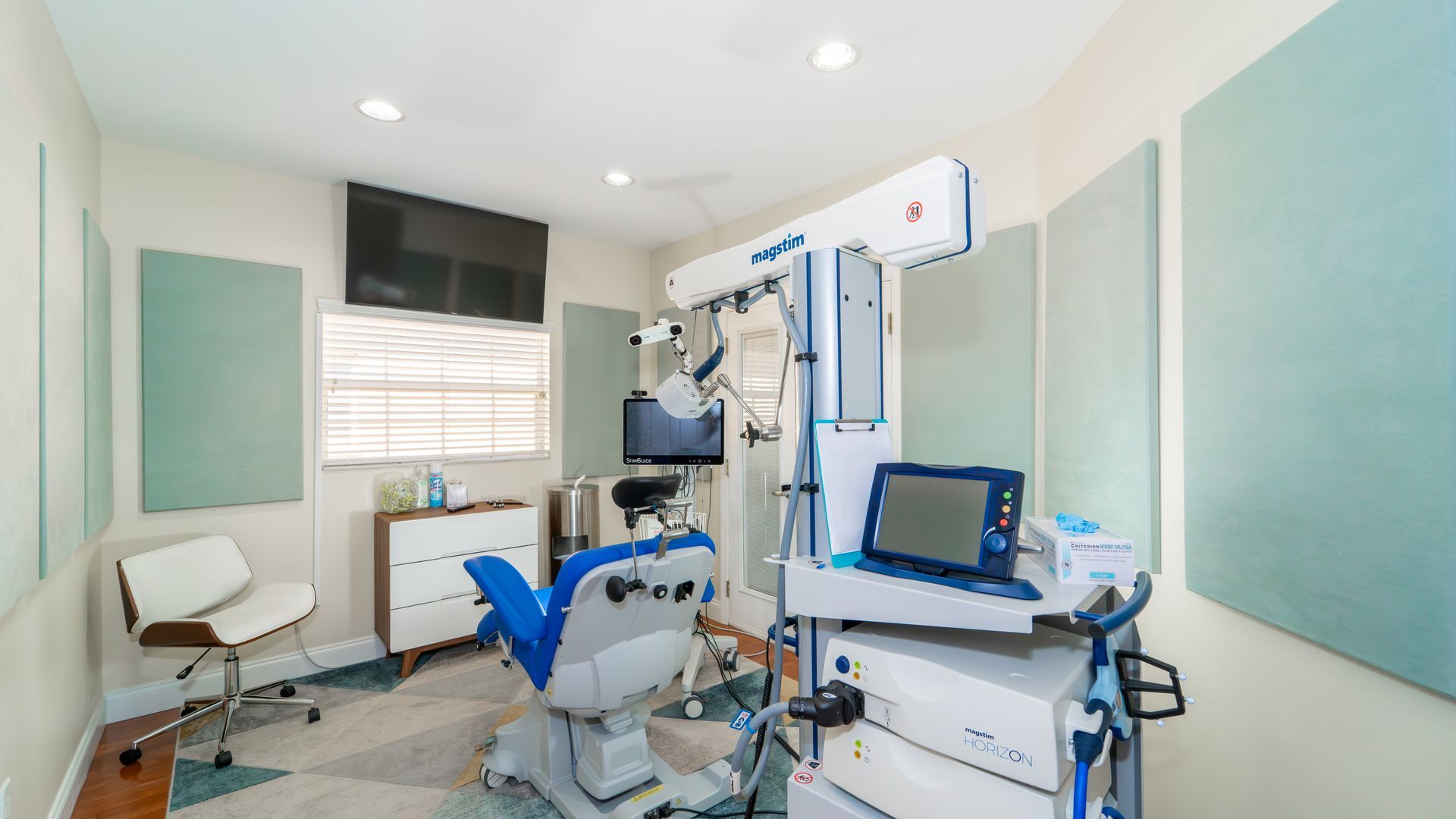 An examination room for an ear specialist, with a blue and white examination chair and medical equipment. Includes neutral-colored walls, sound-dampening panels, and a television.