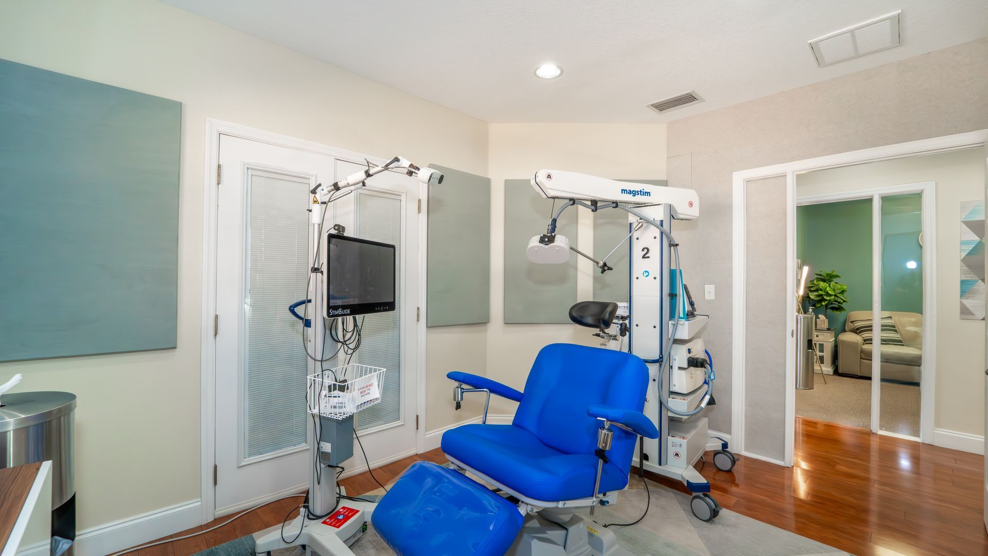A medical exam room features a blue chair and equipment. Another room is visible through a doorway.