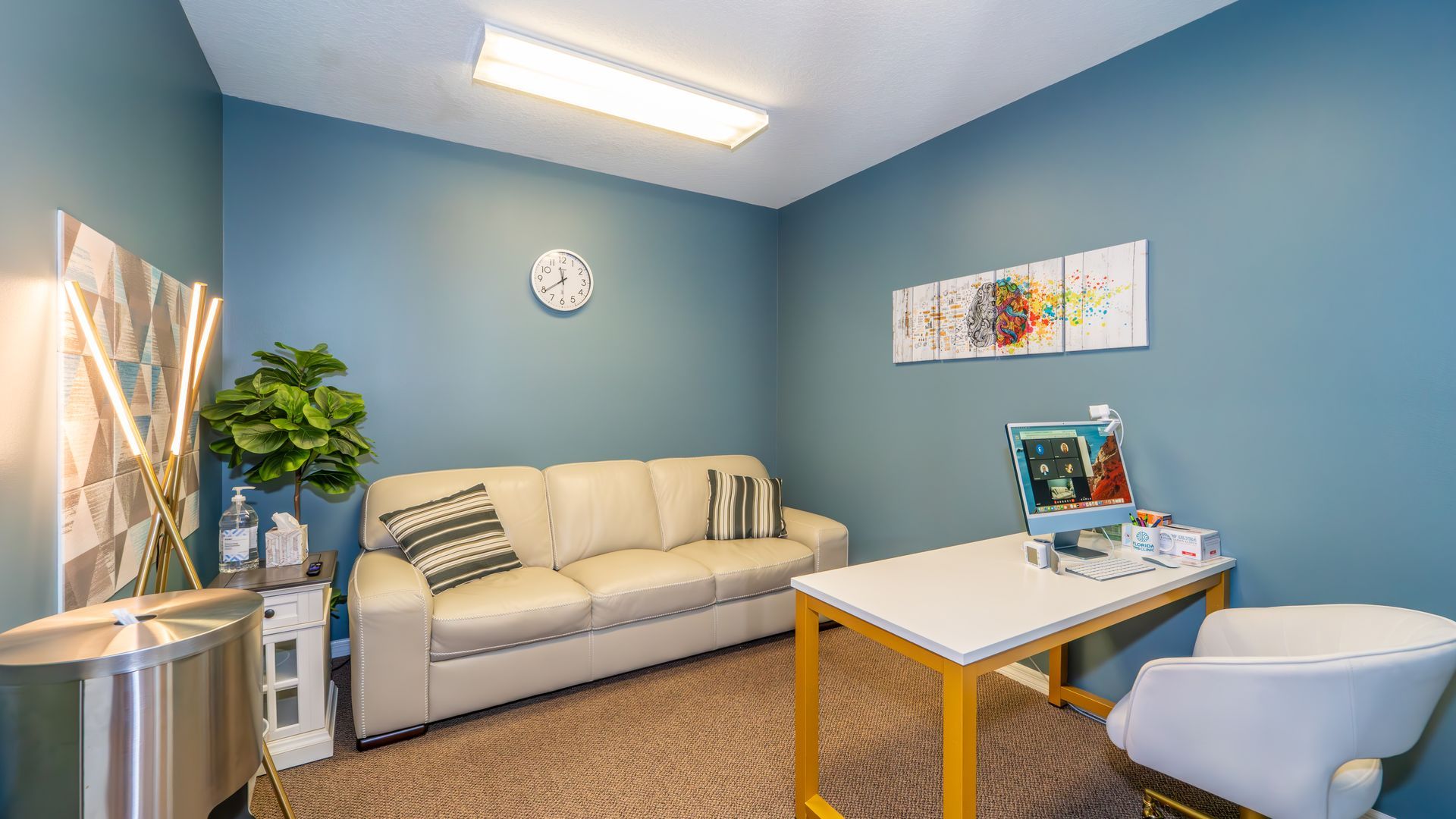 A counseling room with blue walls, a cream-colored sofa, and a desk with a chair. A plant, artwork, and clock are visible.