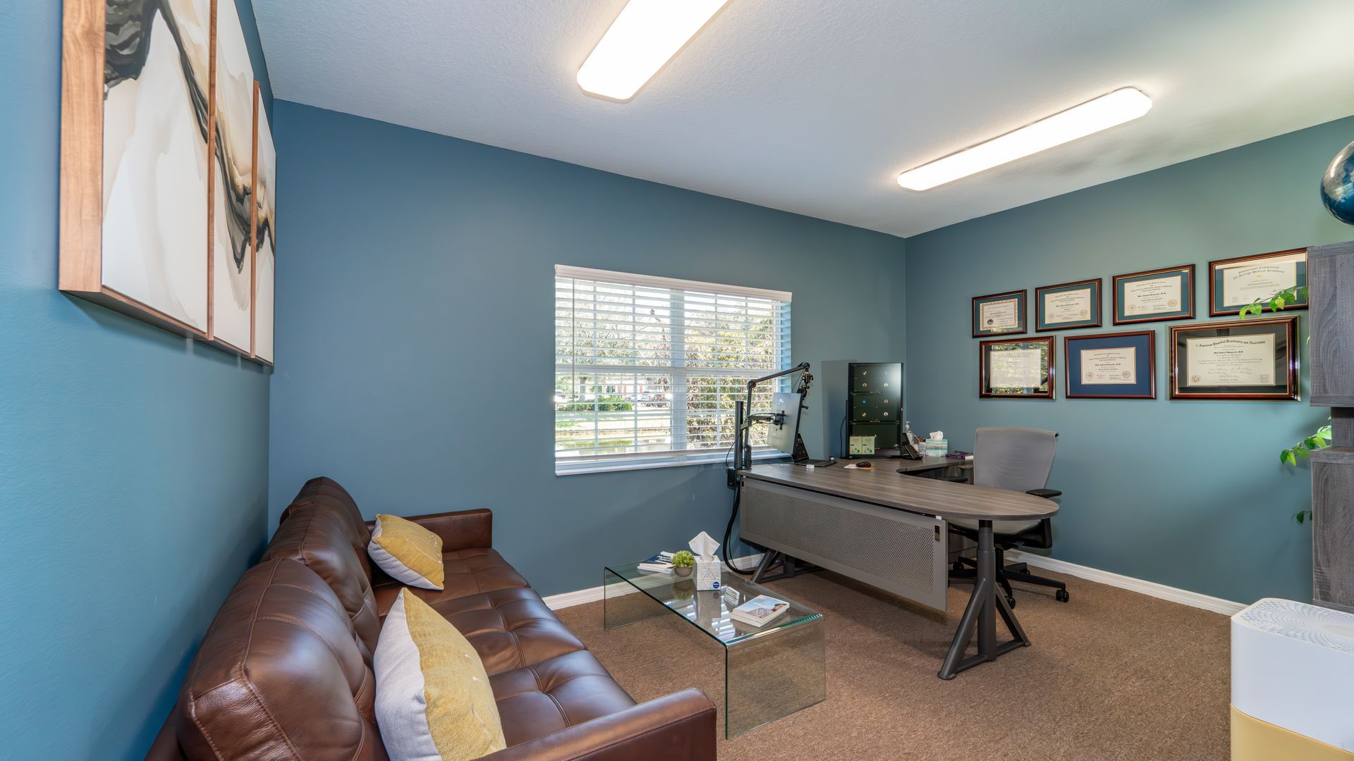 A home office with blue walls, a brown leather sofa, a desk with a computer, and framed diplomas.