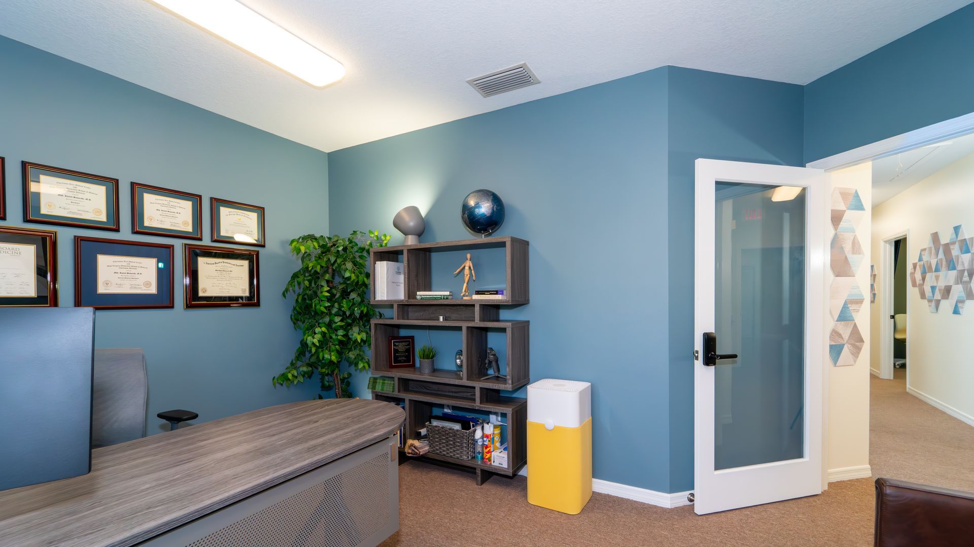 A modern office with blue walls, a desk, shelves, a plant, and a door leading to a hallway.