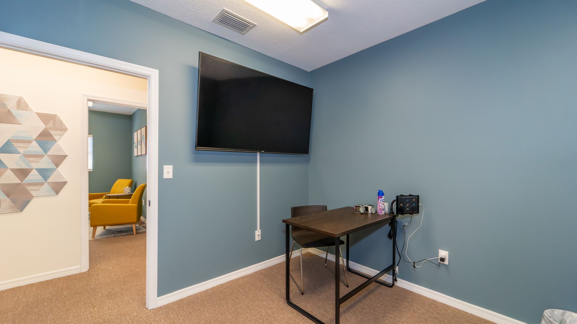 A small office with a blue wall and a mounted TV. A desk with a chair sits below the TV, and another room is visible through a doorway.