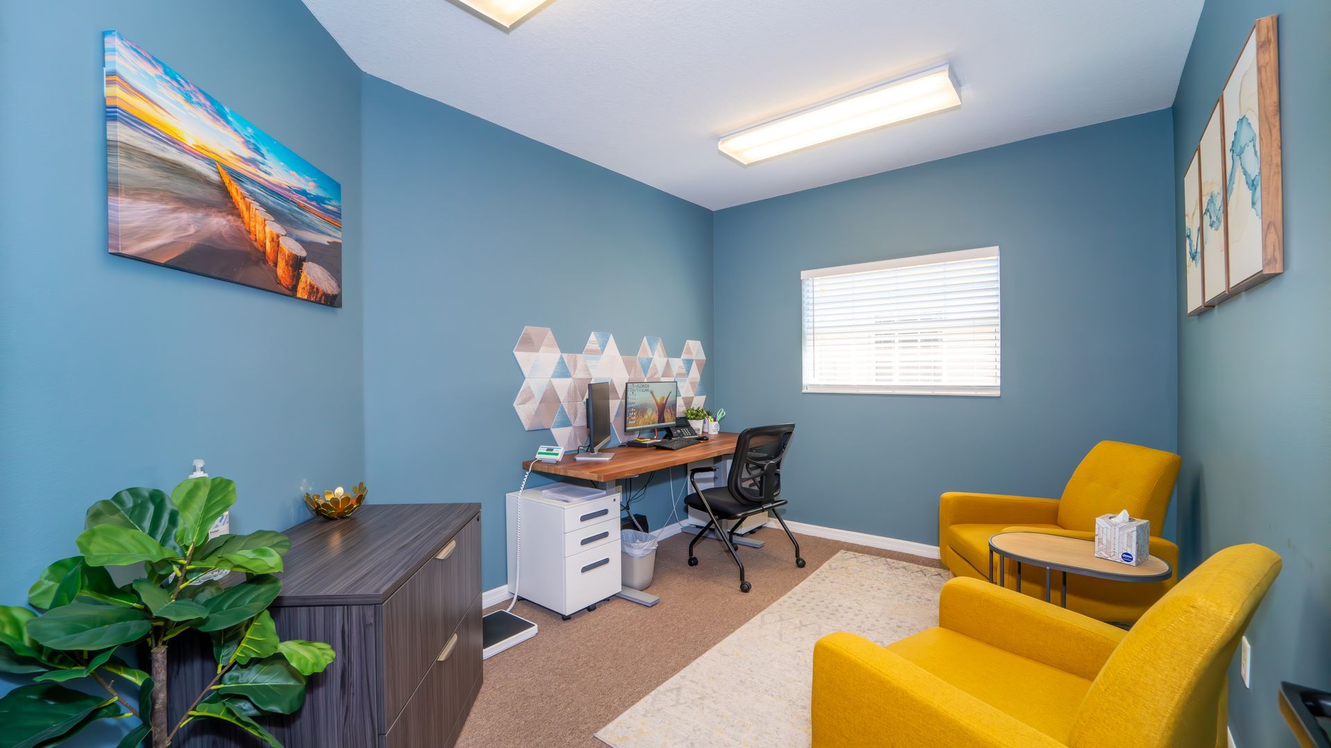 A small office space with blue walls, a desk, yellow armchairs, and decorative art pieces.