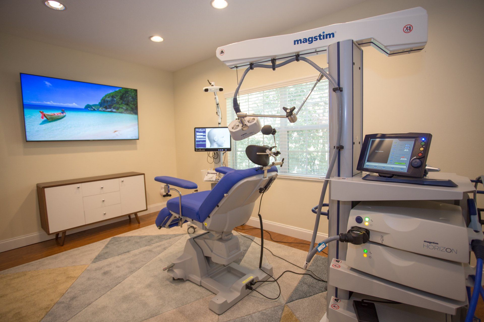 A medical room with a patient chair and machinery. A TV shows a beach scene.
