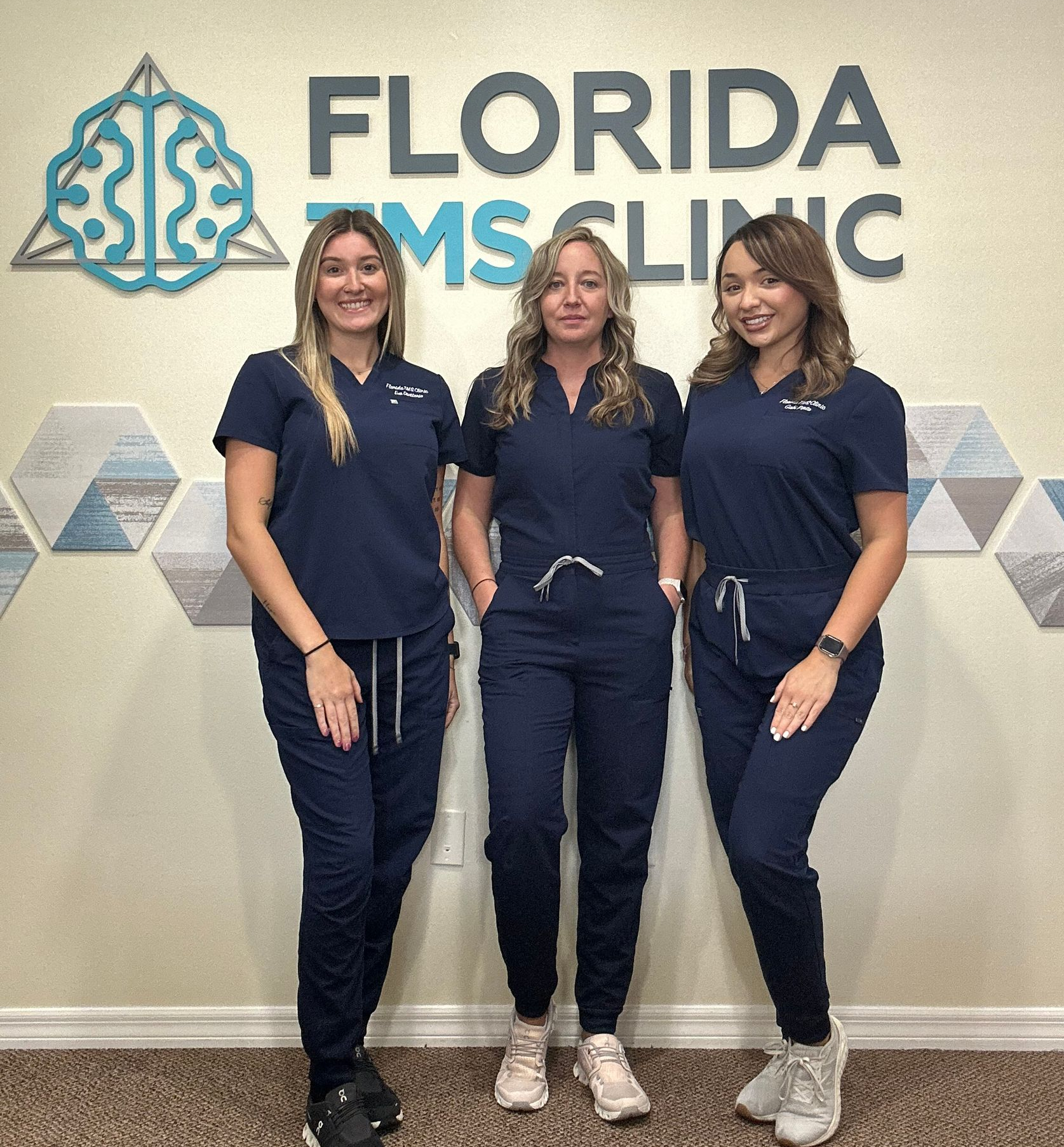 Two women smiling at the front desk of Florida TMS Clinic. They wear blue scrubs. The logo is above them.