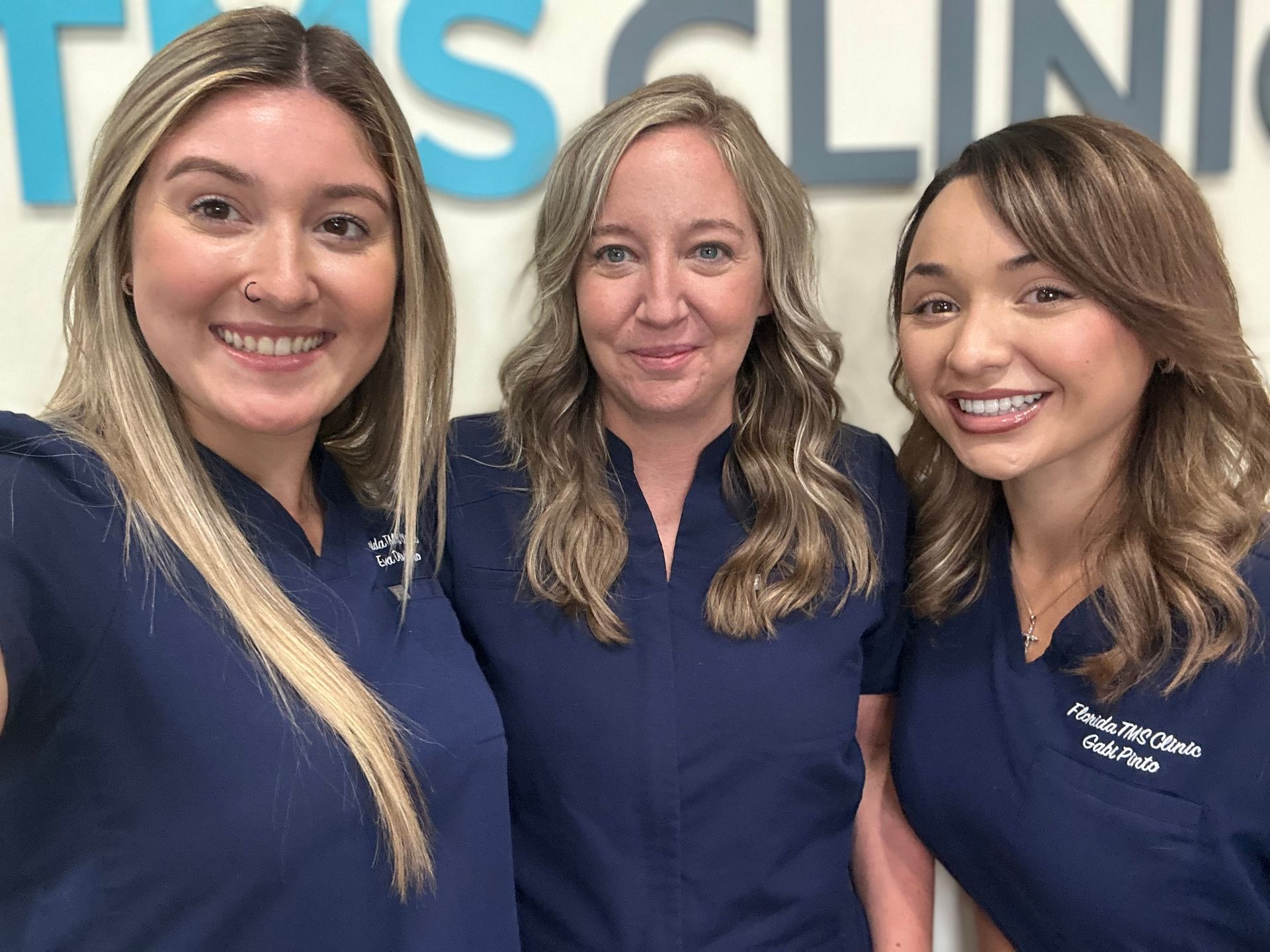 Three women in navy scrubs smiling in front of a clinic sign.