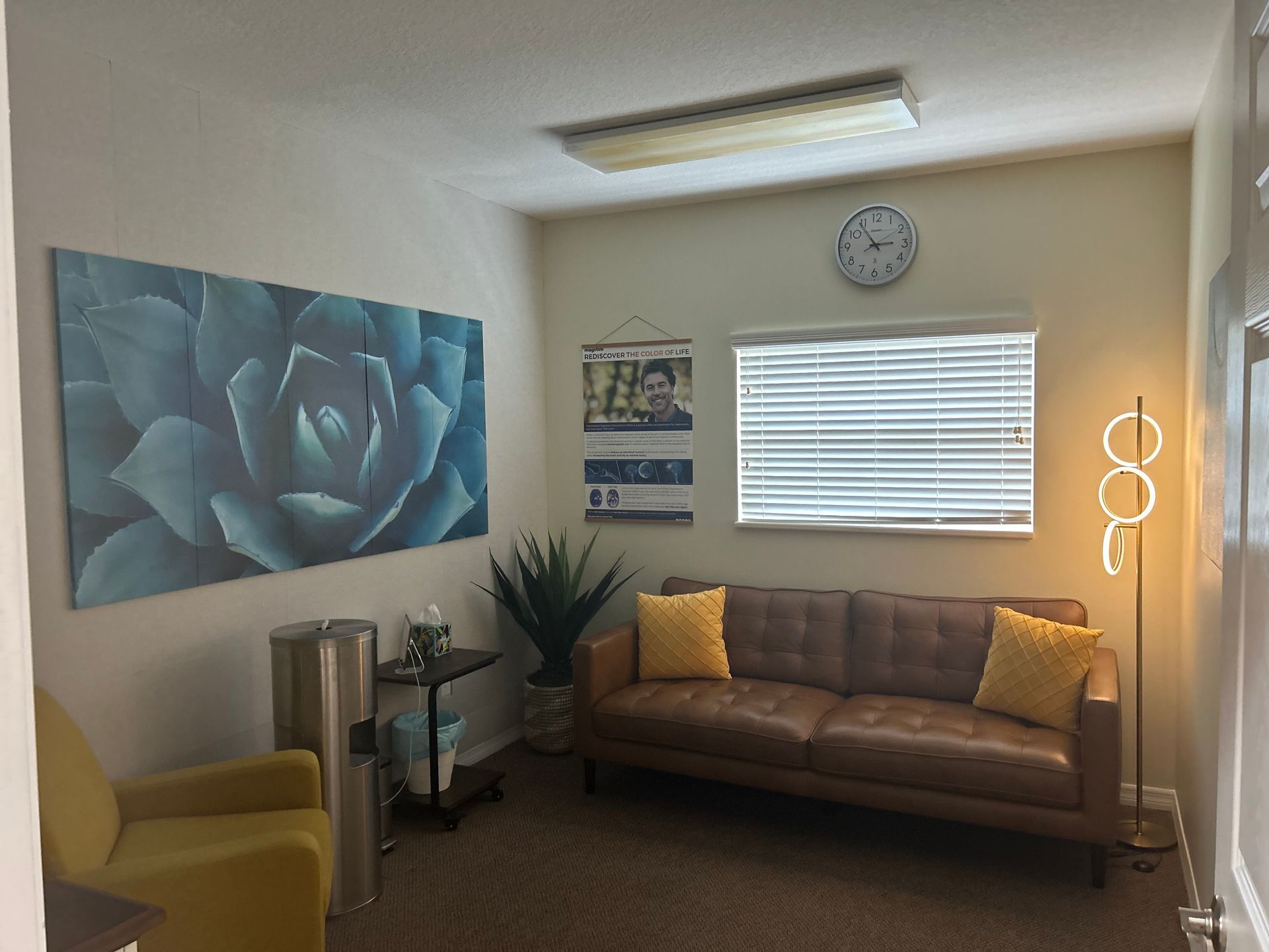 A waiting room with a brown sofa, blue floral art, a clock, and a golden lamp.