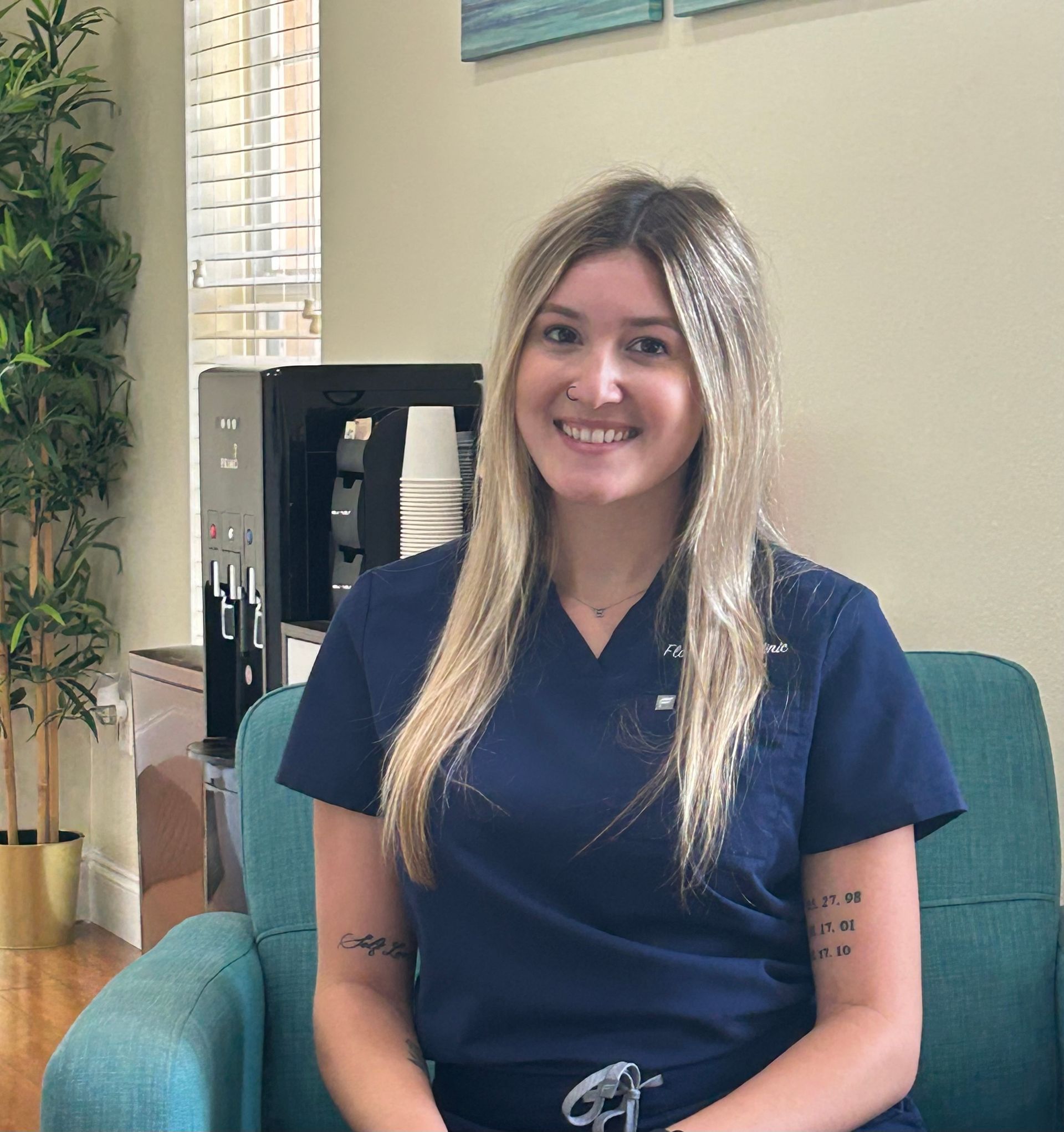 Woman with blonde hair in blue scrubs smiles, seated in a teal chair.