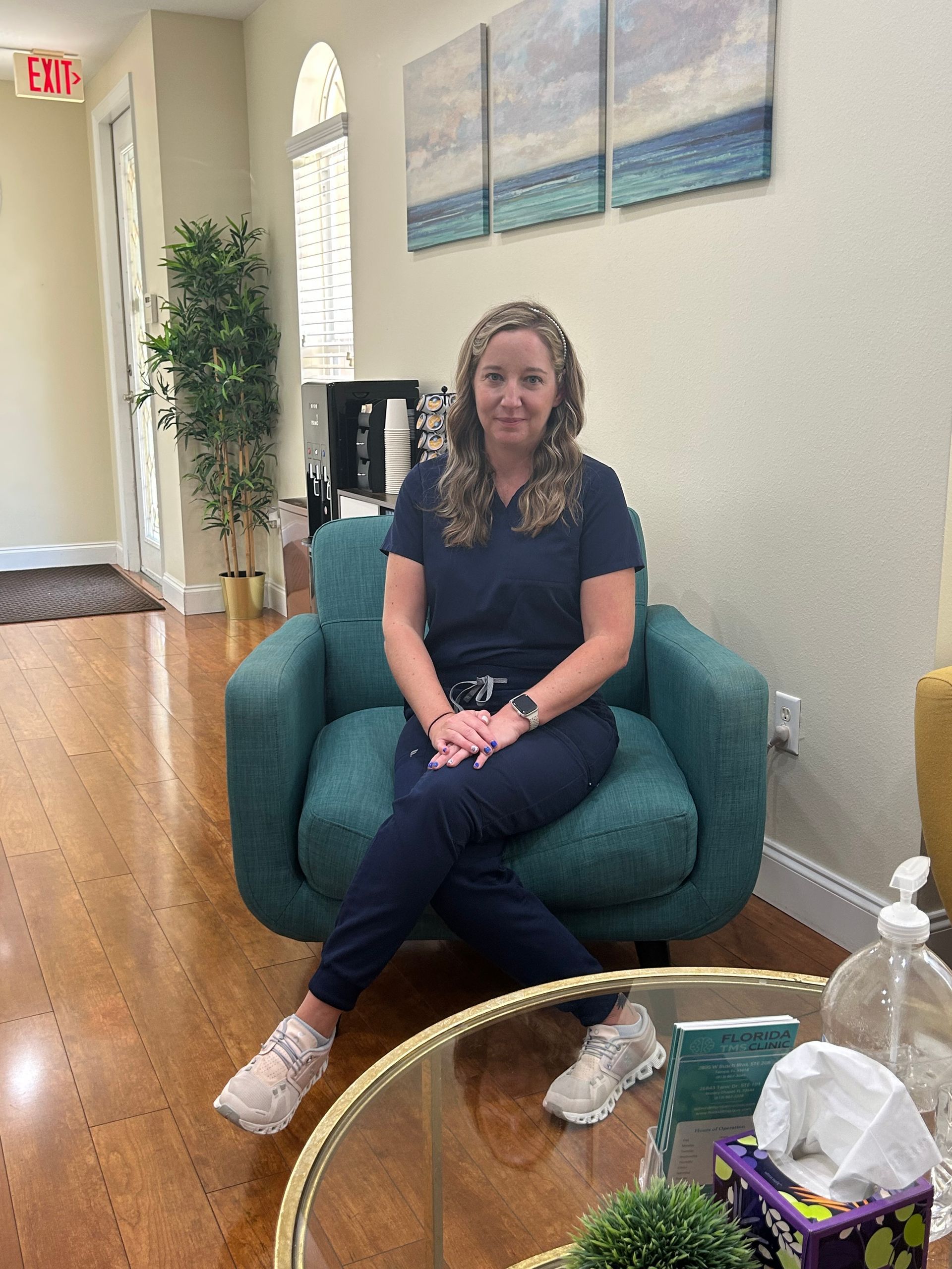Woman in blue scrubs sitting in teal chair, smiling. Interior, art on wall.