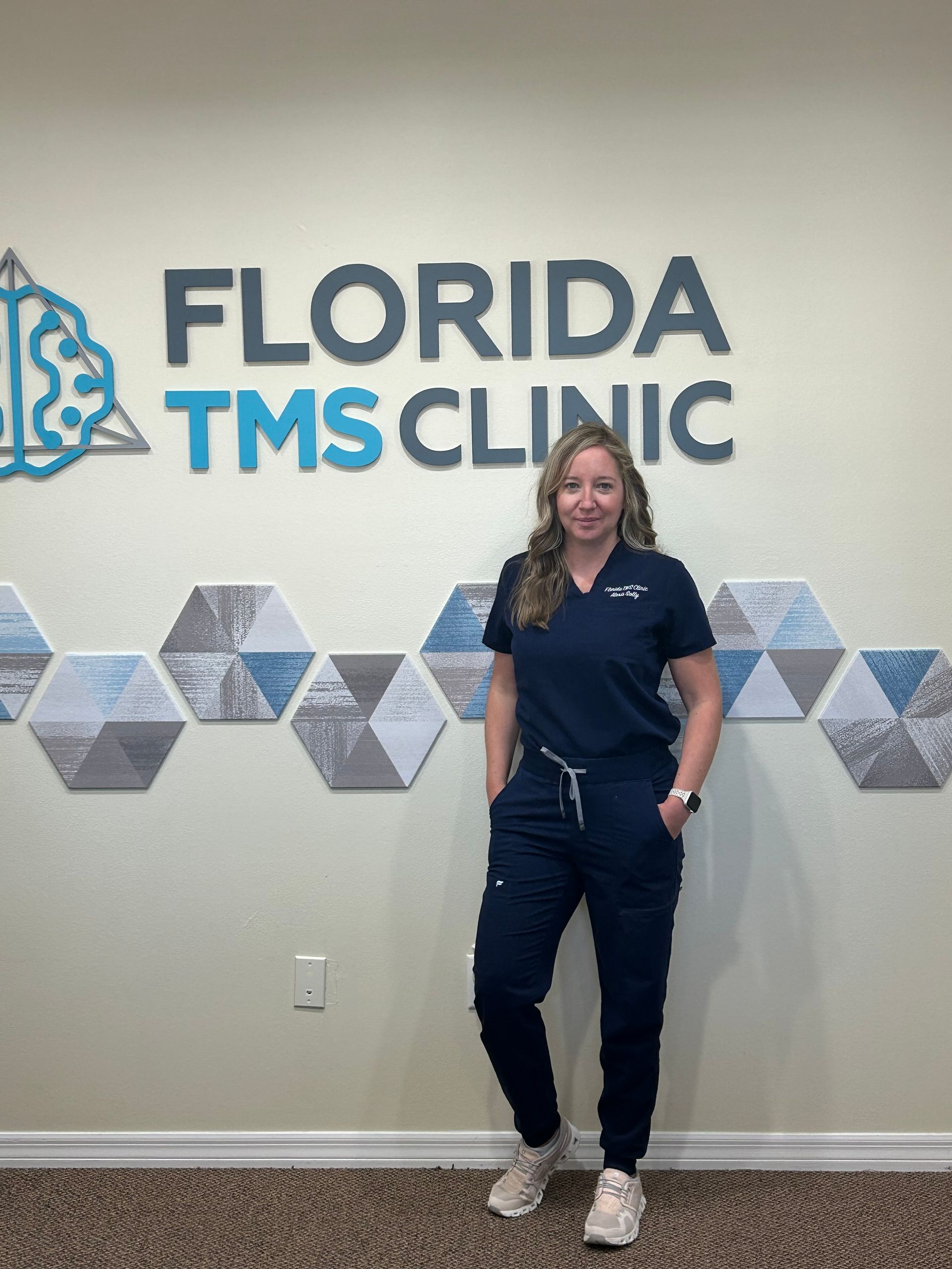 Woman in navy scrubs stands in front of Florida TMS Clinic sign, hands in pockets.