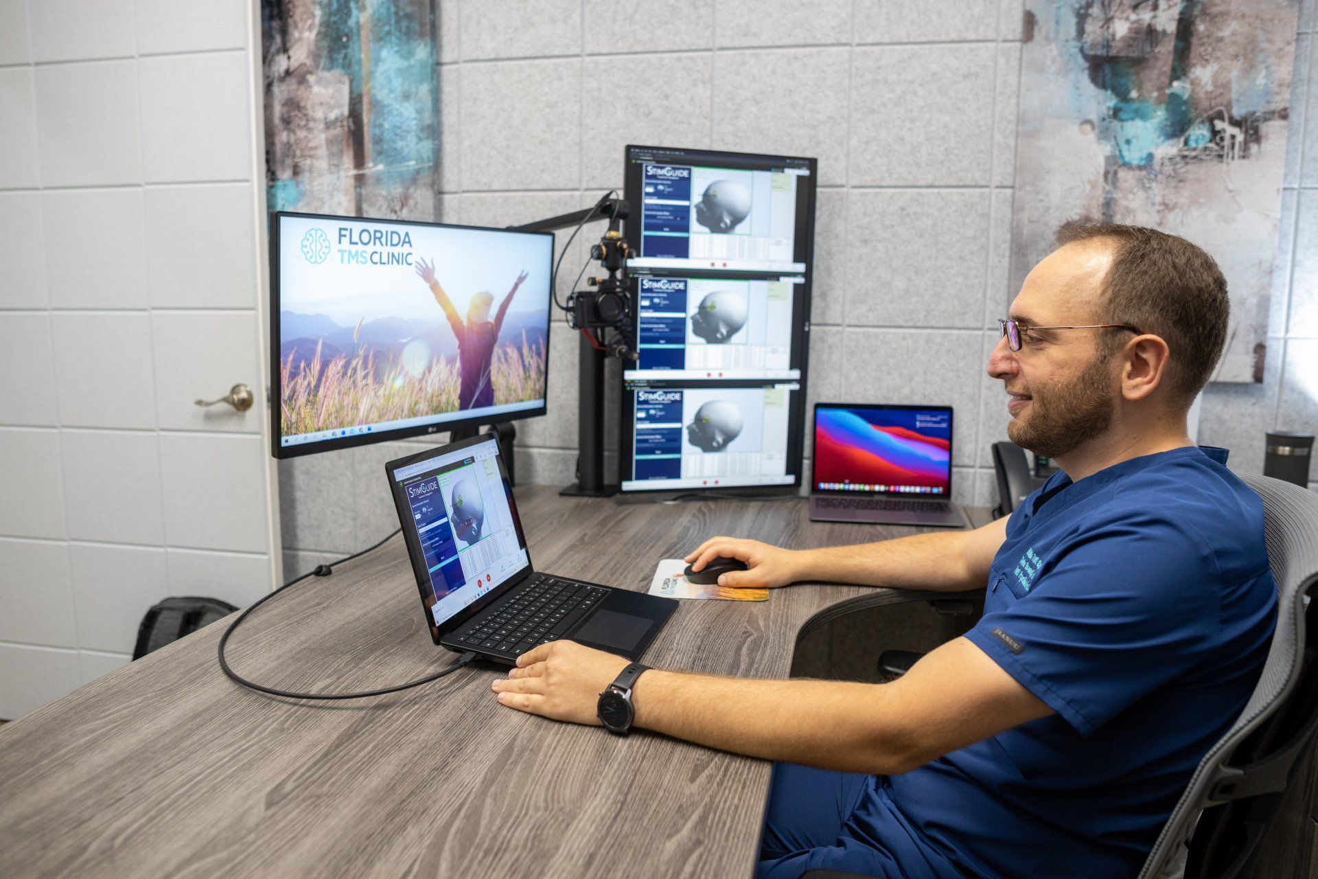 Man in blue scrubs sits at a desk with multiple monitors. He smiles while using a laptop and mouse, looking at the screens.