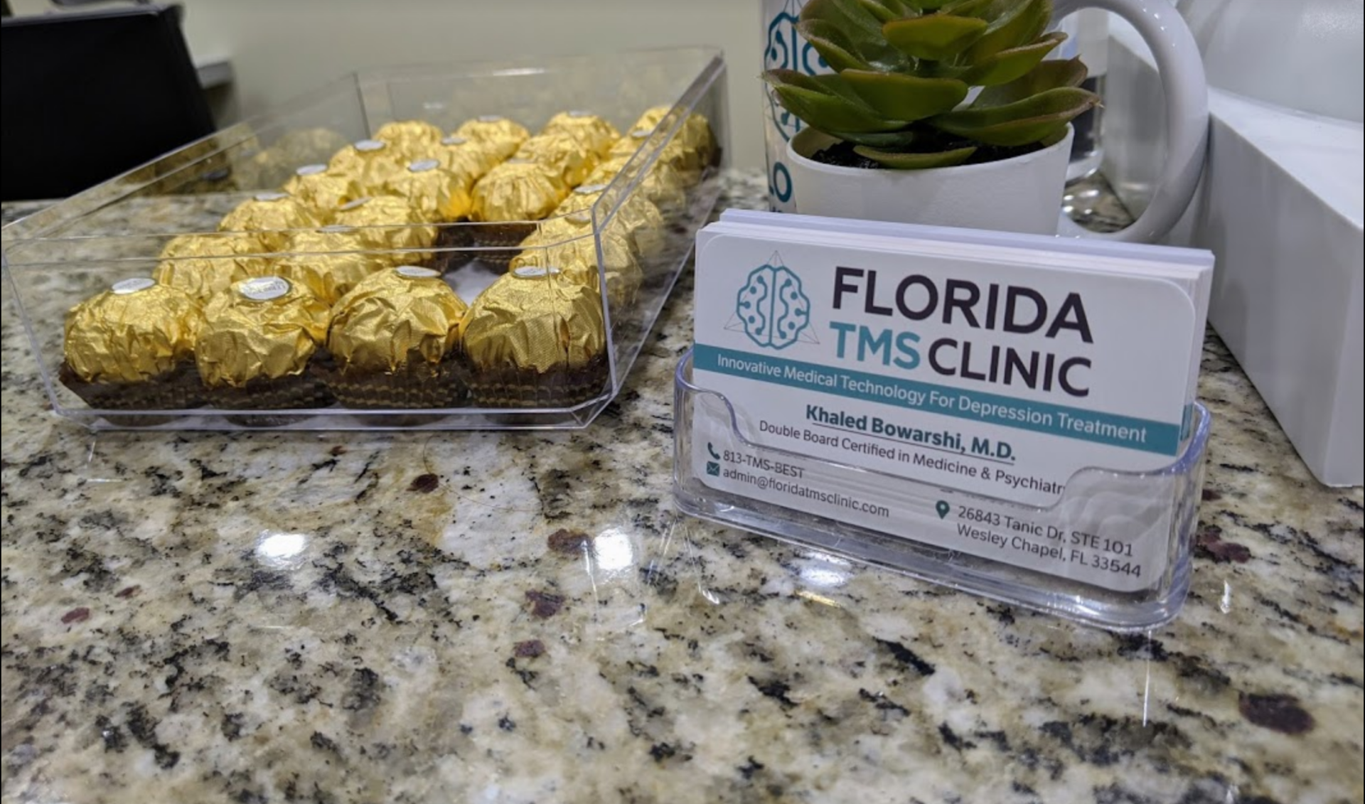 A clear plastic holder displays business cards for Florida TMS Clinic. A tray of gold-wrapped chocolates sits to the left on a marble surface.