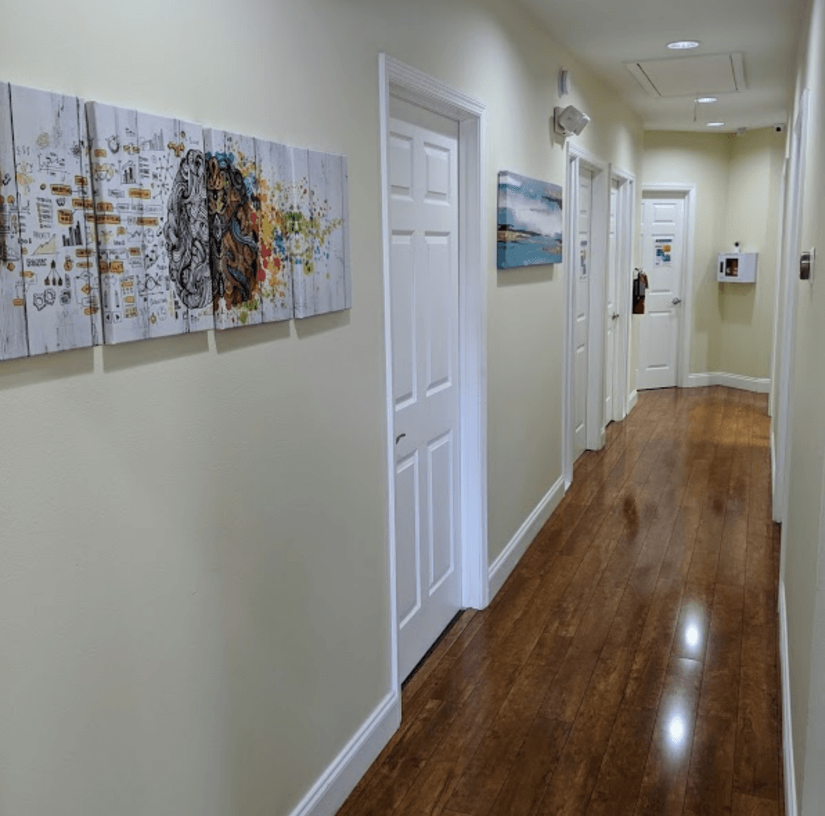 Long, bright hallway with hardwood floors, several closed white doors, and abstract art on the walls.