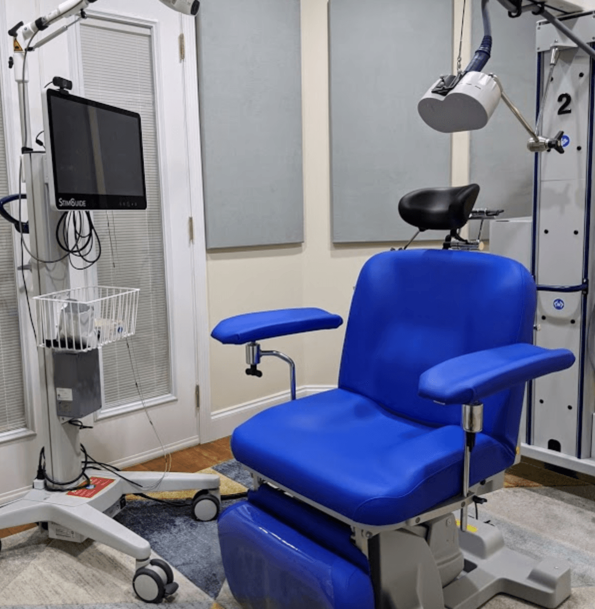 Blue medical chair with armrests, near a white equipment cart with a monitor and a white light. The setting appears to be a medical examination room.