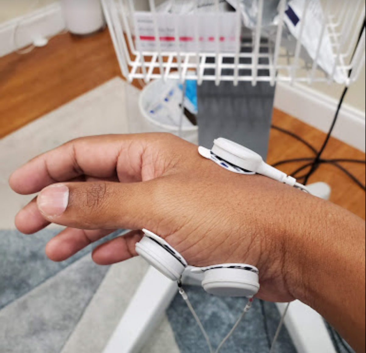 A person's hand with white electrodes attached, likely for medical testing. The hand rests on a light gray surface with a basket and wood floor in the background.