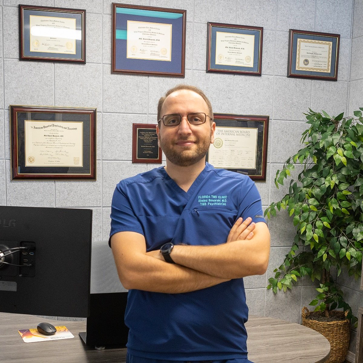 A man with a beard and glasses in blue scrubs stands in an office with arms crossed, diplomas on the wall.