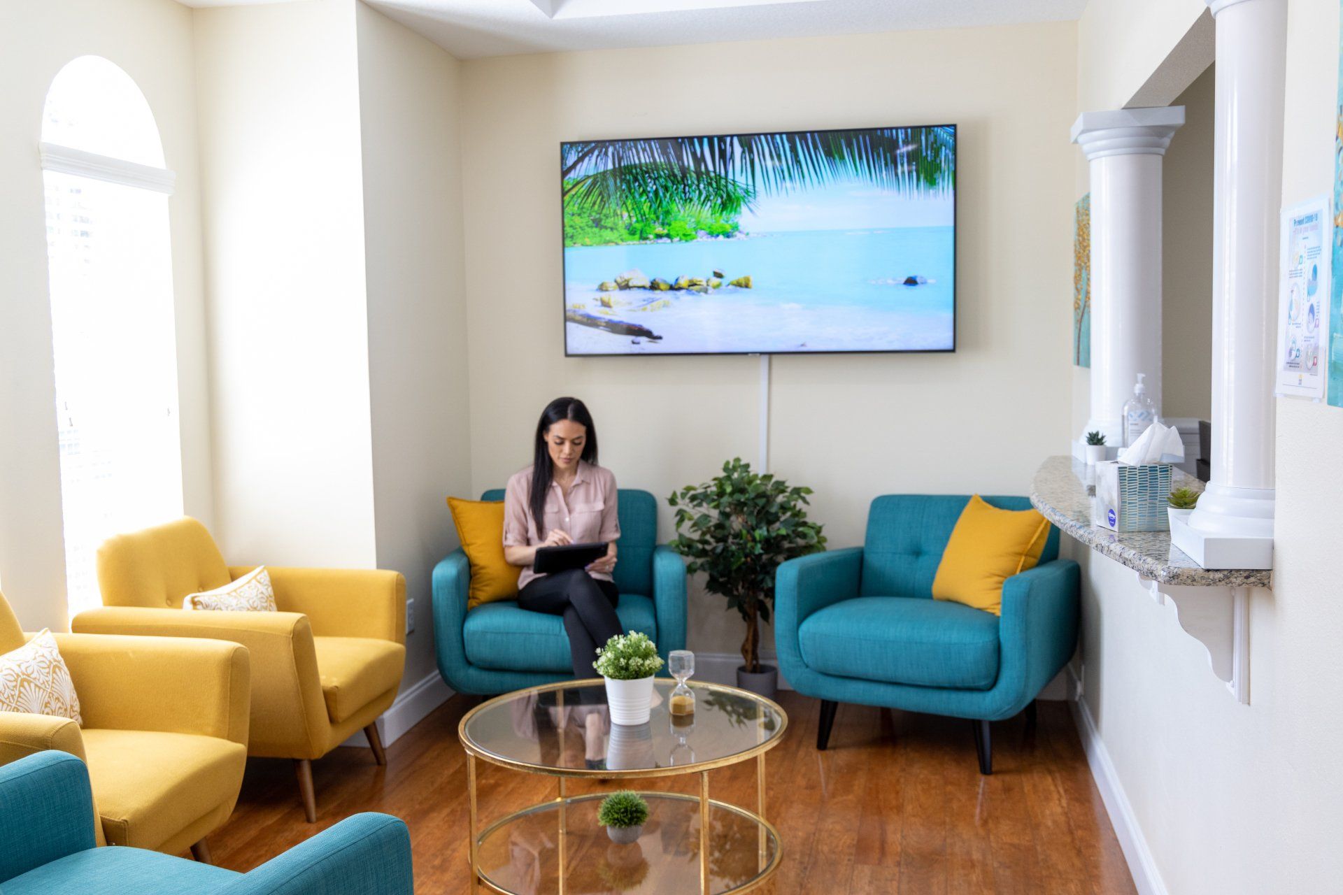 Woman in a waiting area reads a tablet, surrounded by yellow and teal armchairs, a TV displaying a tropical scene, and a round coffee table.
