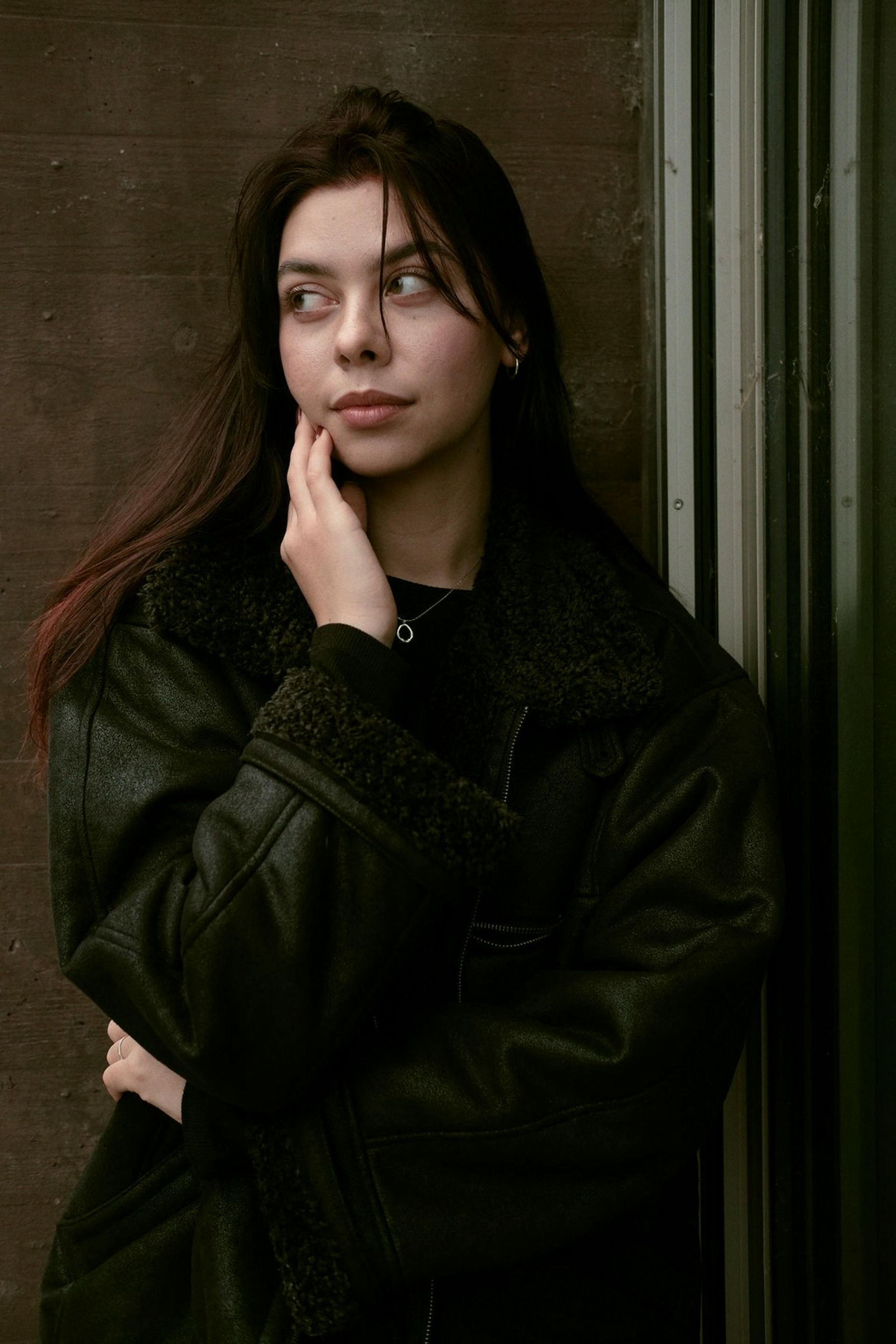 Woman in black coat leans against a dark wall, hand to her cheek, looking to the side.