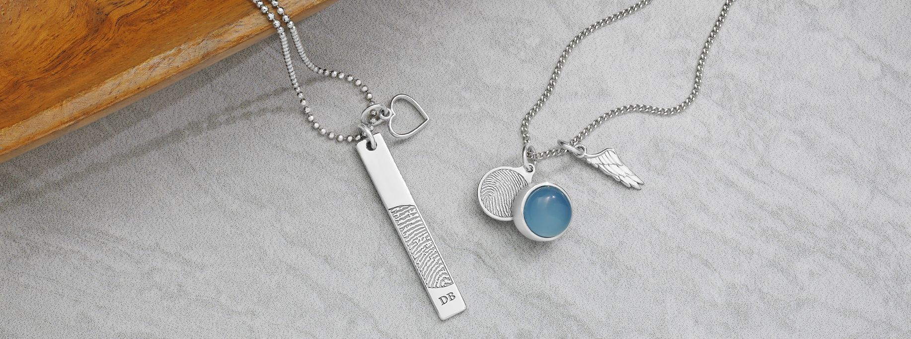 Two silver necklaces with pendants on a gray surface, one with a bar, the other with a blue gem.