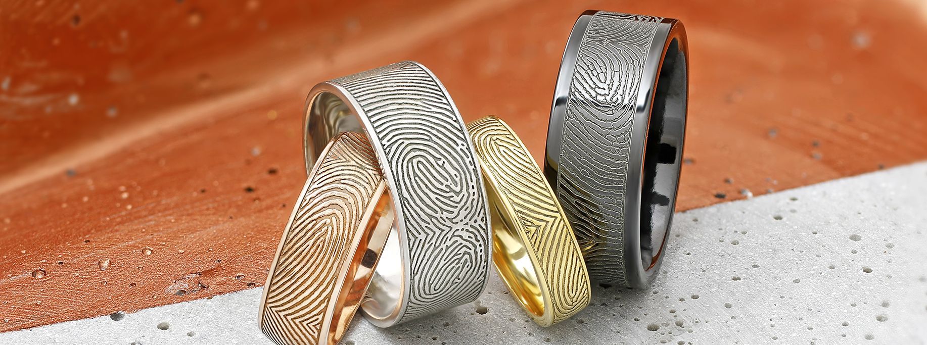 Close-up of four patterned metal rings, gold, silver, and black, resting on a textured orange and gray surface.