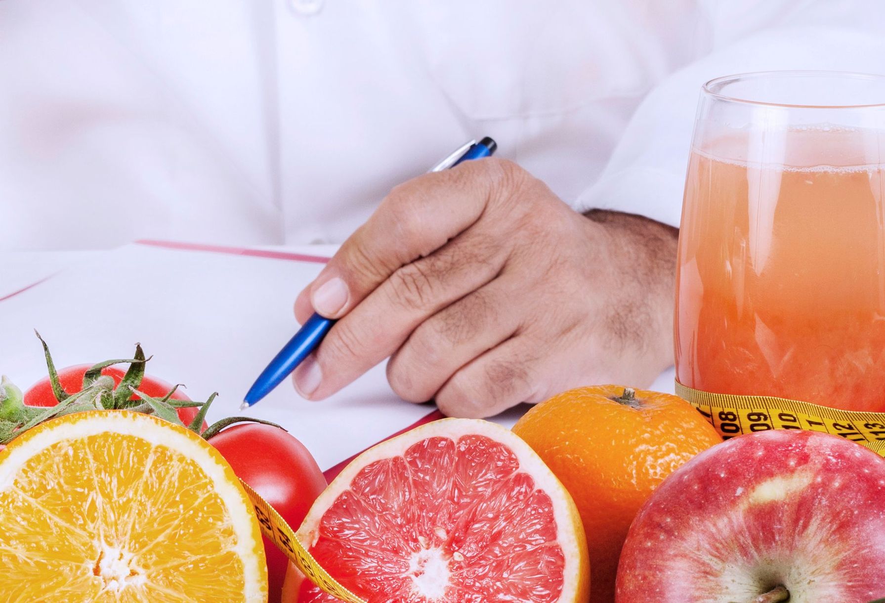 Persona in camice bianco che scrive su un foglio, circondata da frutta e un bicchiere di succo.