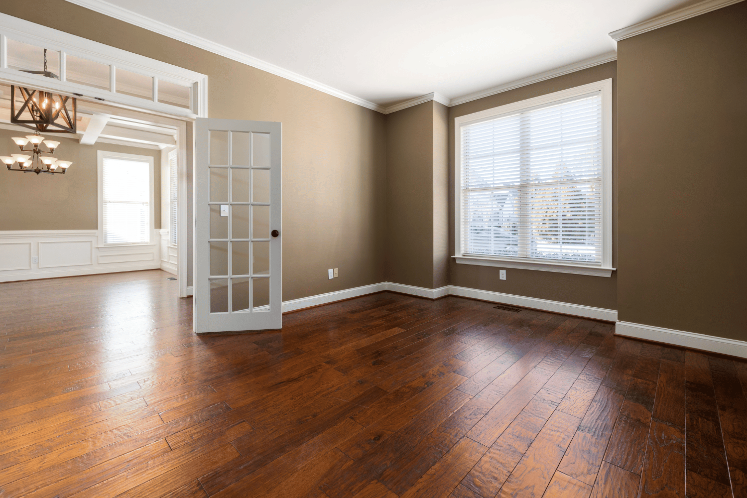 An empty room with dark wood flooring, tan walls, a white French door, and a large window looking into an adjacent room.