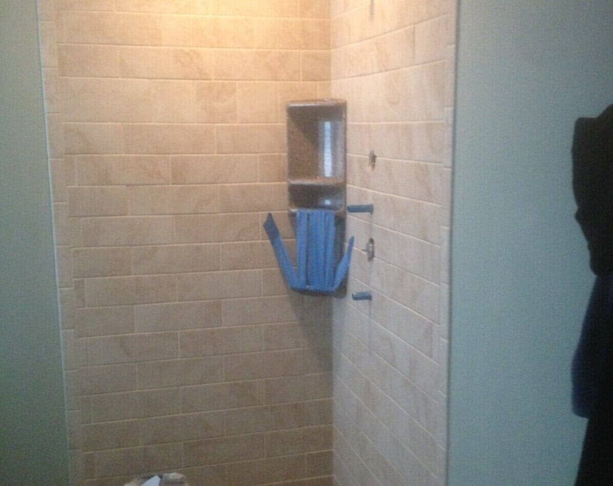 A tiled shower corner featuring a recessed wall shelf containing a blue plastic organizer and a mounted shower handle.