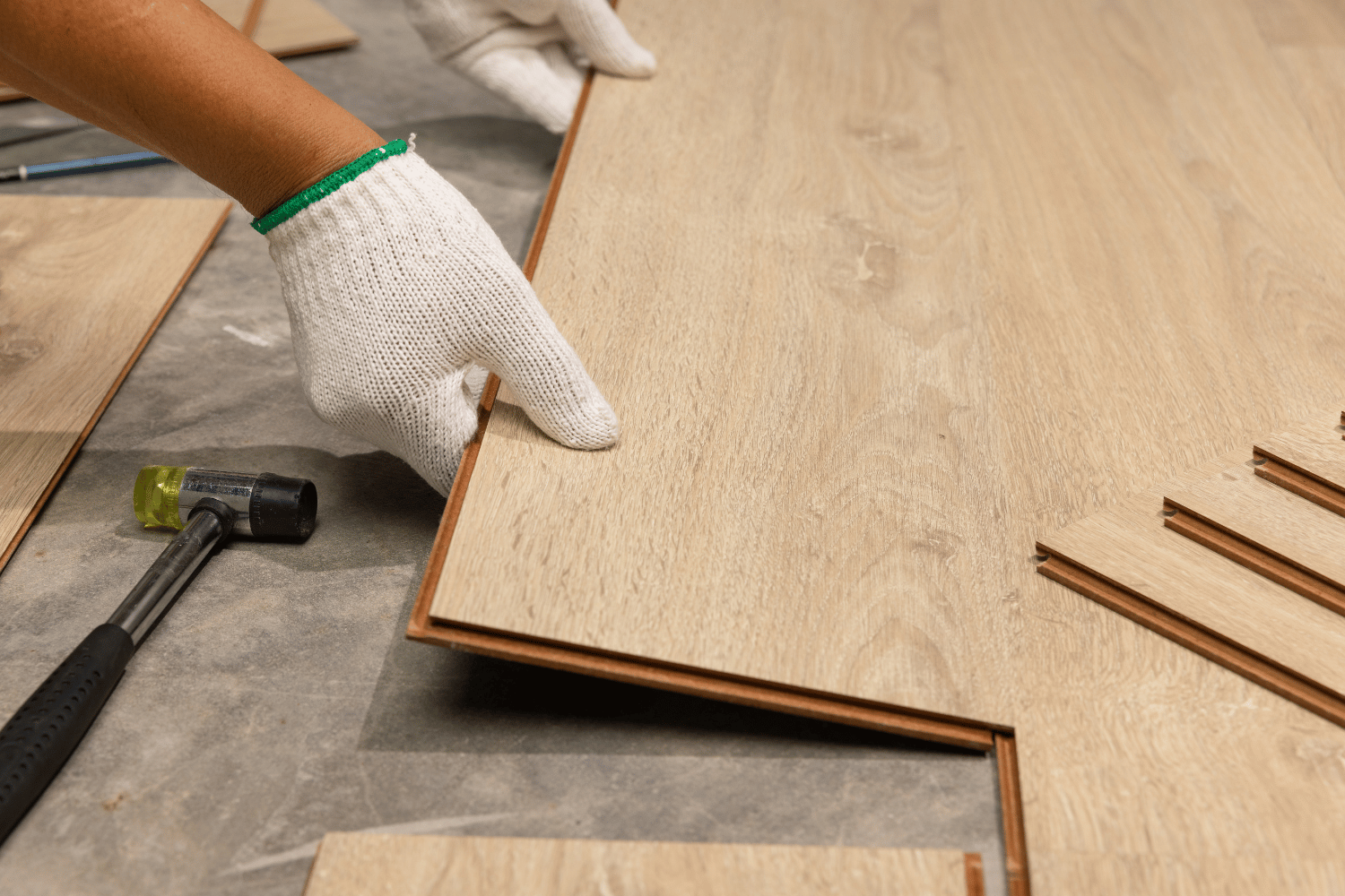 Hands in white gloves align a wood-look laminate floor plank next to a mallet on a construction site.