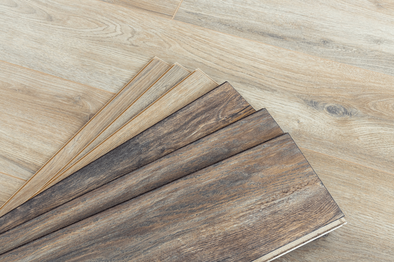 A fan of vinyl flooring samples ranging from light oak to dark, weathered wood tones resting on a matching plank floor.