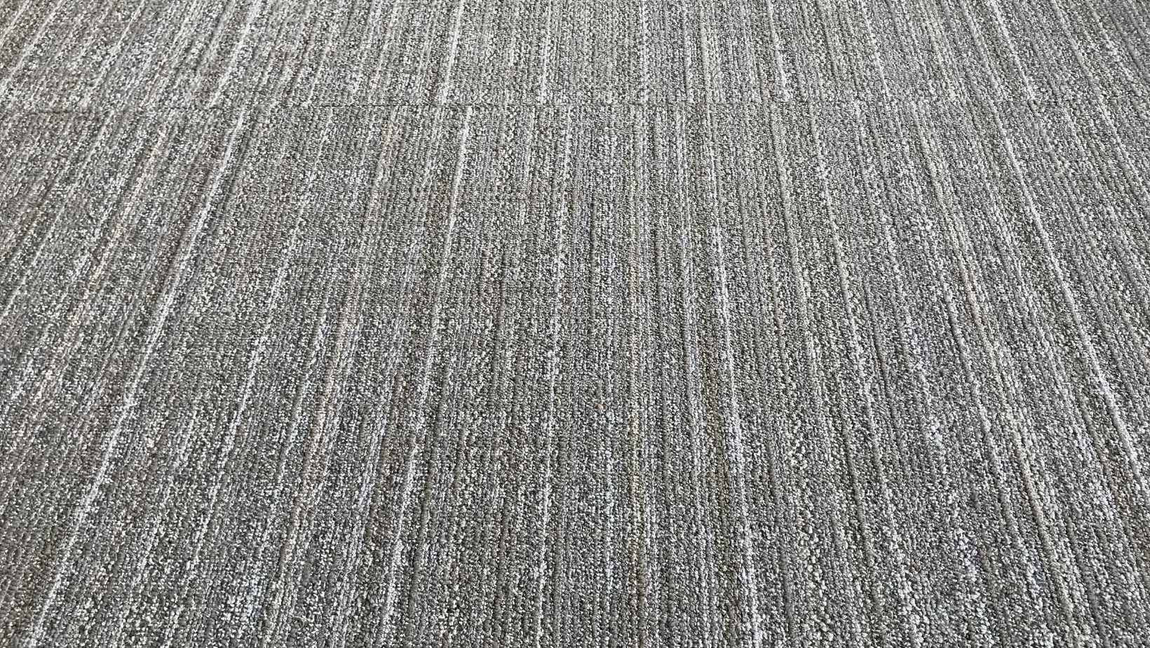 A close-up view of textured gray carpet tiles with a linear, vertical pattern.