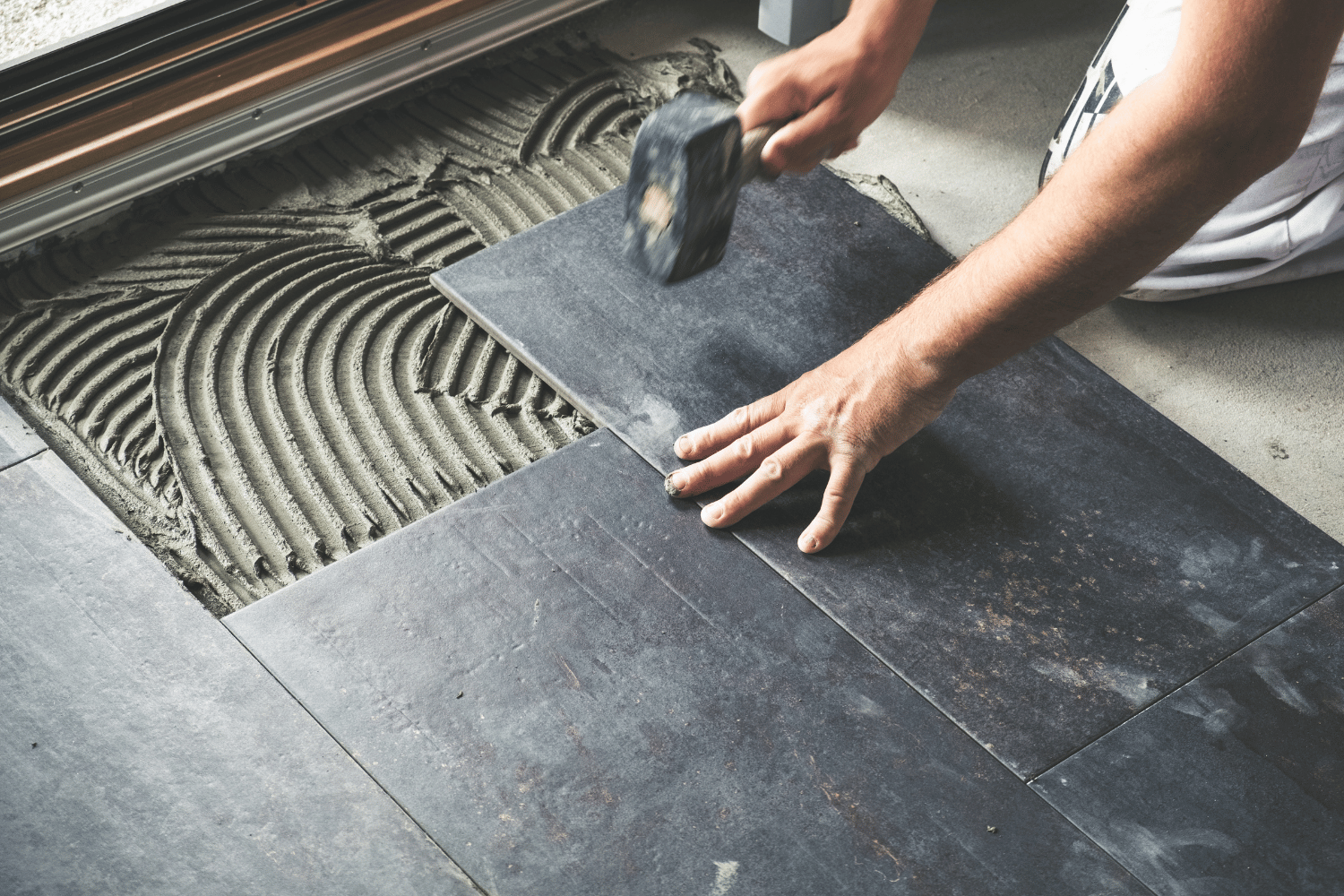 A person uses a rubber mallet to secure a dark gray tile onto a bed of adhesive mortar.