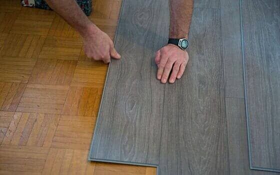A pair of hands installs grey wood-look laminate flooring panels over an existing parquet floor.
