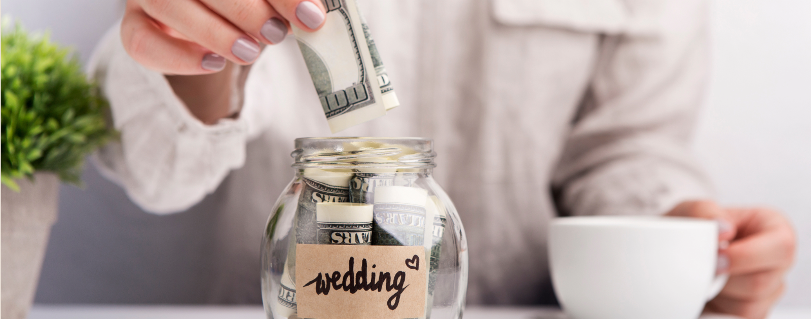 Woman's hand putting a one hundred dollar bill into a glass jar of money with the label Wedding on it.