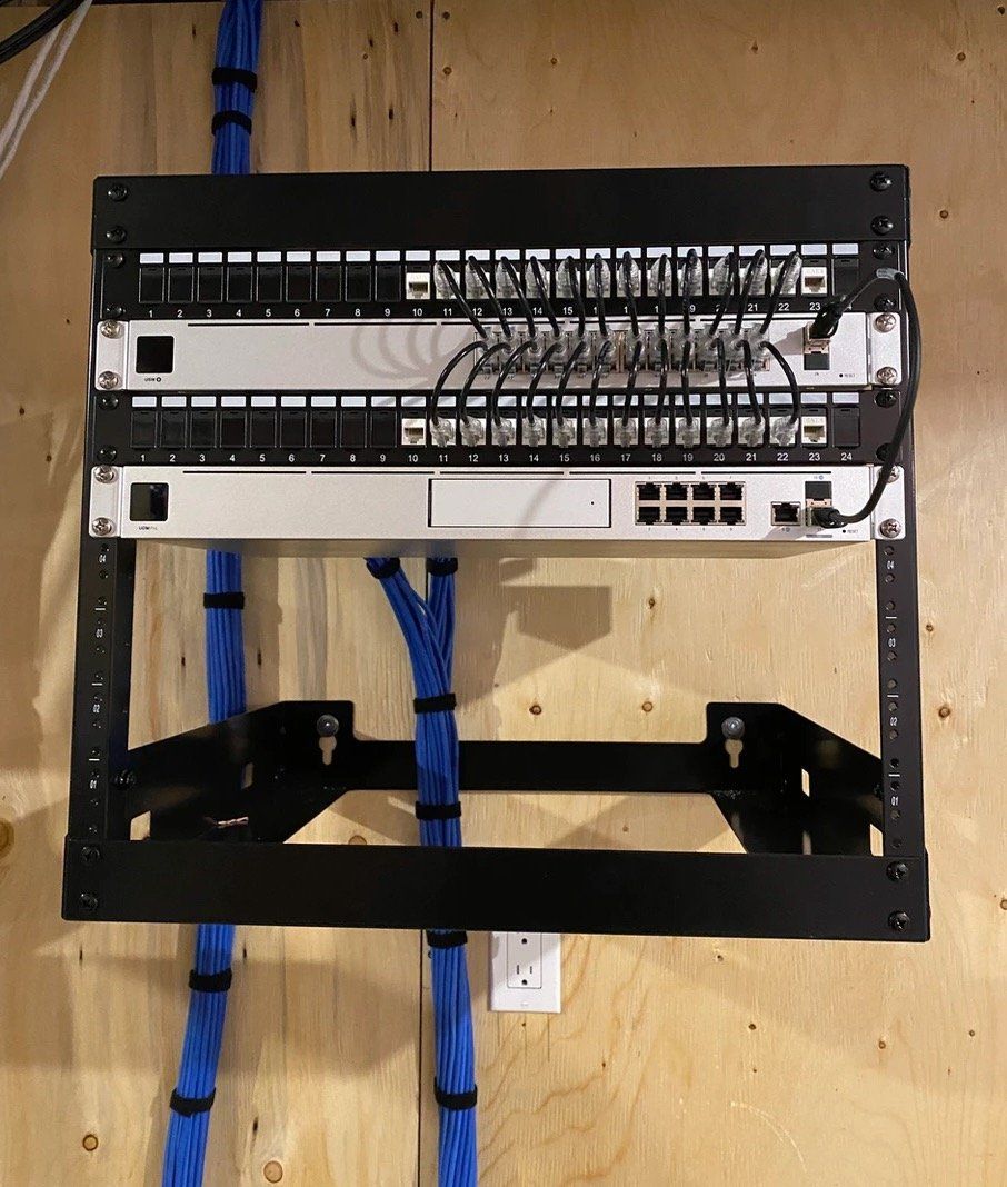 Black network rack on a wooden wall with patch panel, switch, and blue network cables.