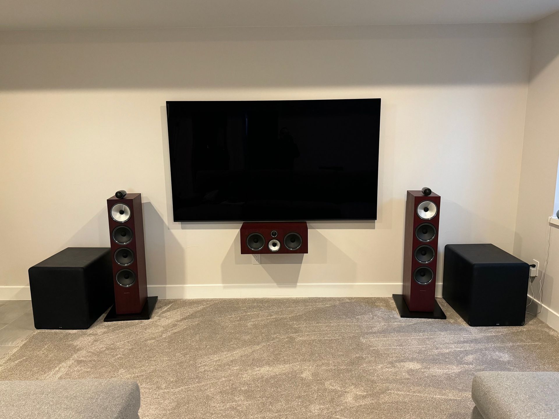 A home theater setup: large TV, speakers, subwoofers, and center channel speaker on a tan carpet.