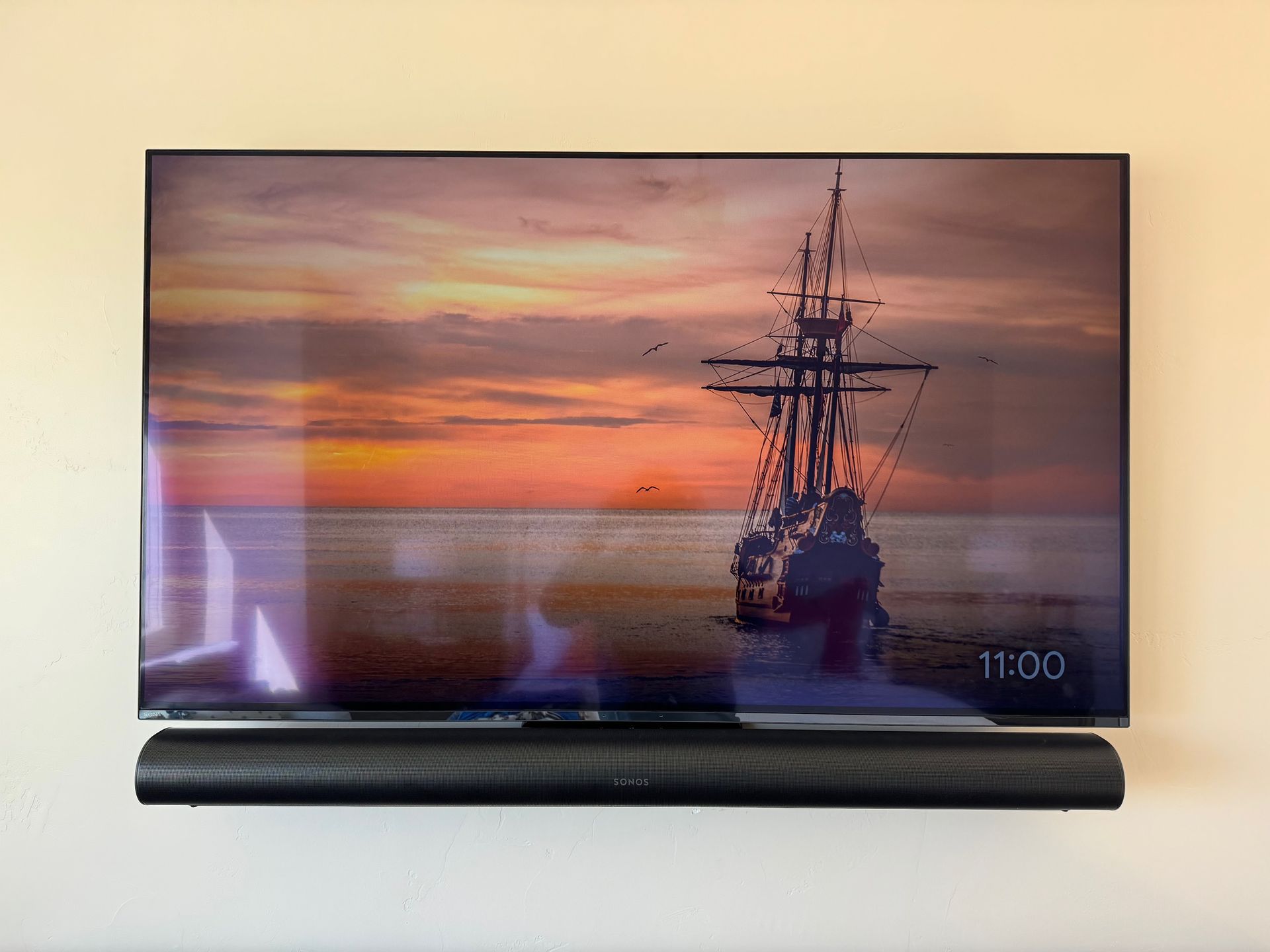 TV displaying a sunset scene with a ship, mounted above a black soundbar on a beige wall.
