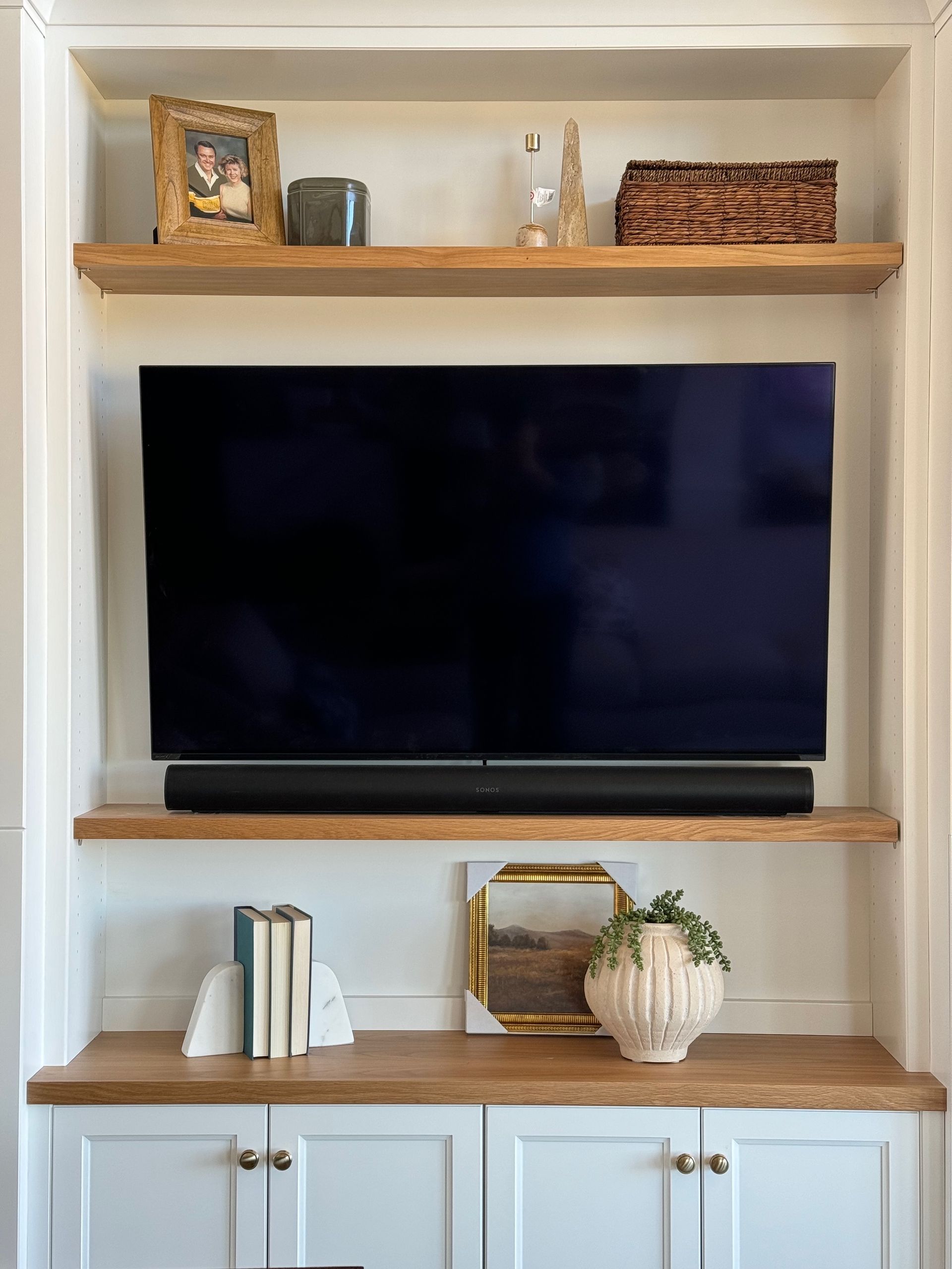 Built-in white cabinet with wood shelves; TV, soundbar, decor (books, picture frame, plant) displayed.